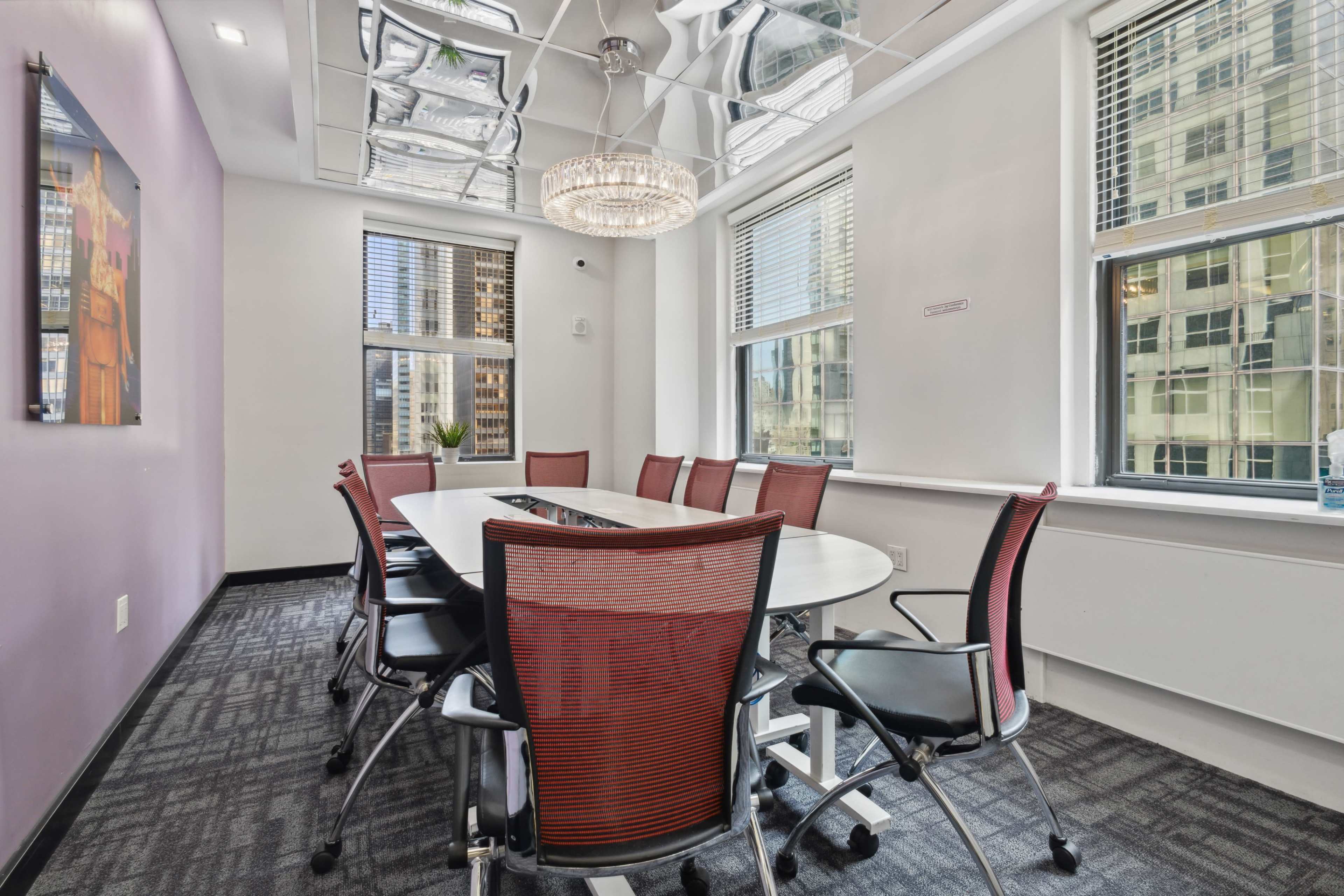 A modern conference room with a large oval table surrounded by red mesh chairs, featuring a chandelier and large windows overlooking a cityscape.