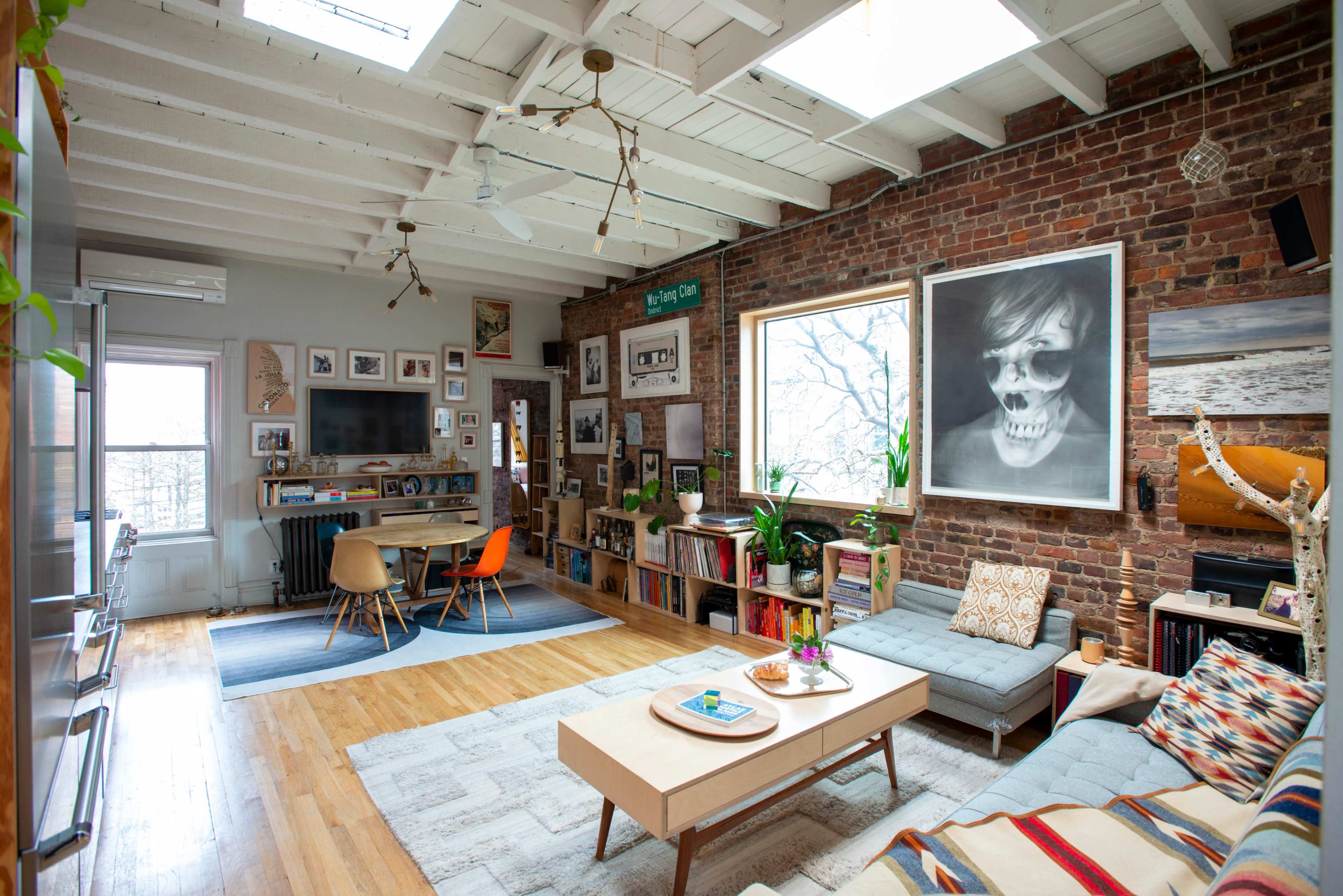 The image shows a stylish living room with exposed brick walls, wooden floors, and a mix of modern and eclectic decor, including a large artwork and a cozy seating area.