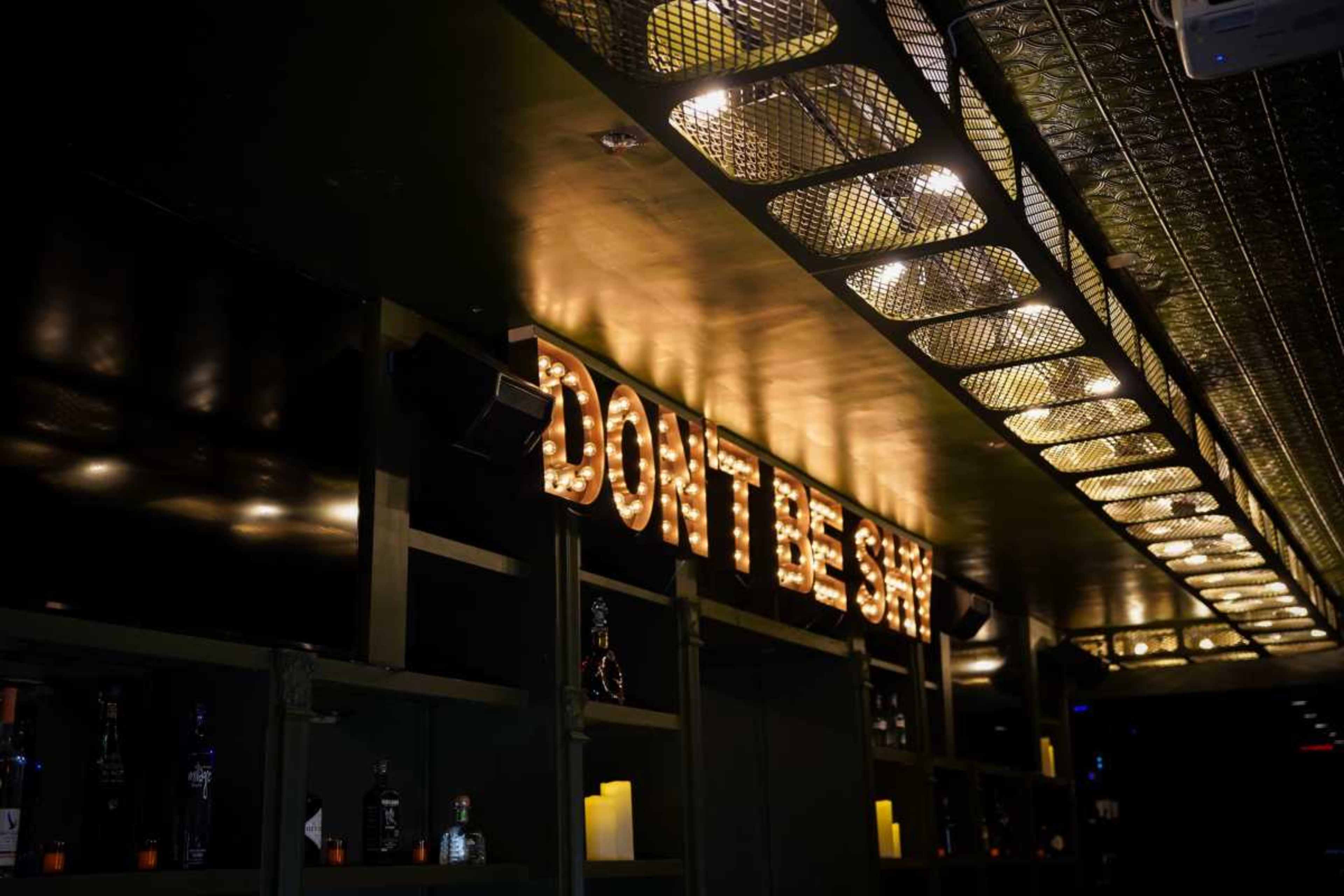 A bar interior features a sign that reads "DON'T BE SHY" illuminated by vintage light bulbs.