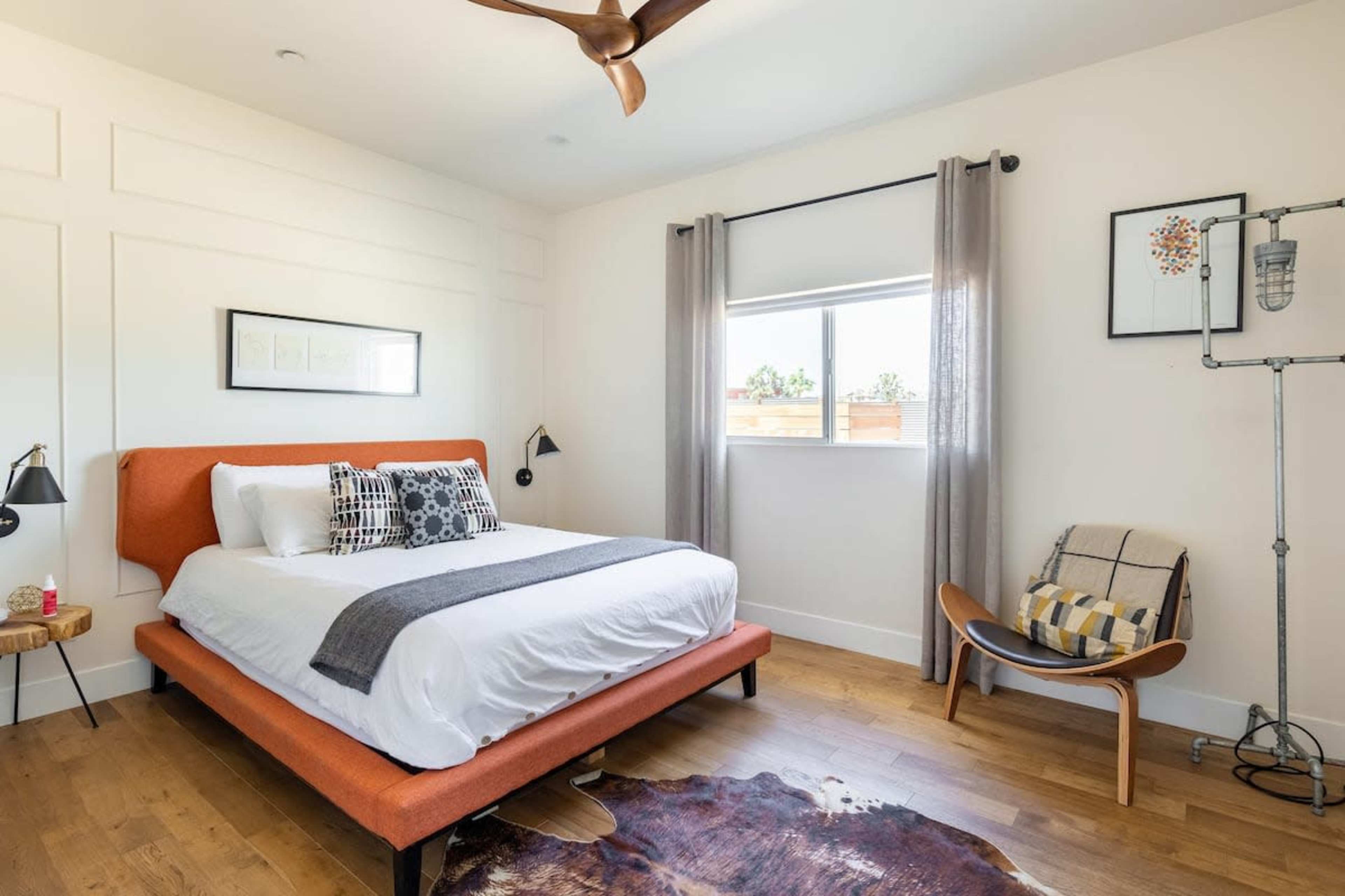 A modern bedroom with an orange bedframe, a white bedspread, and wooden flooring, featuring a window with gray curtains and a small seating area.