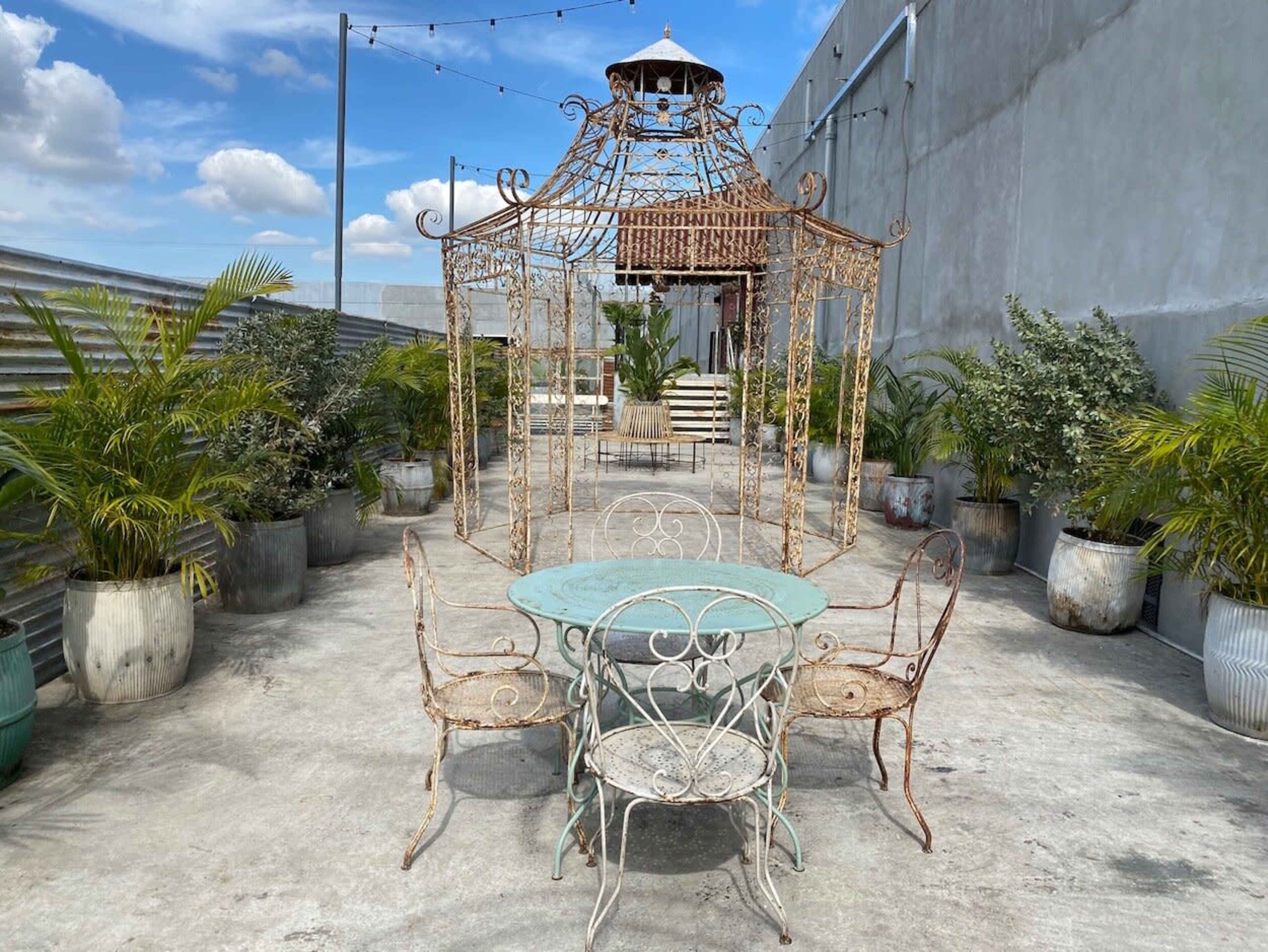 A wrought iron gazebo with a circular table and two chairs is situated on a concrete terrace surrounded by potted plants.