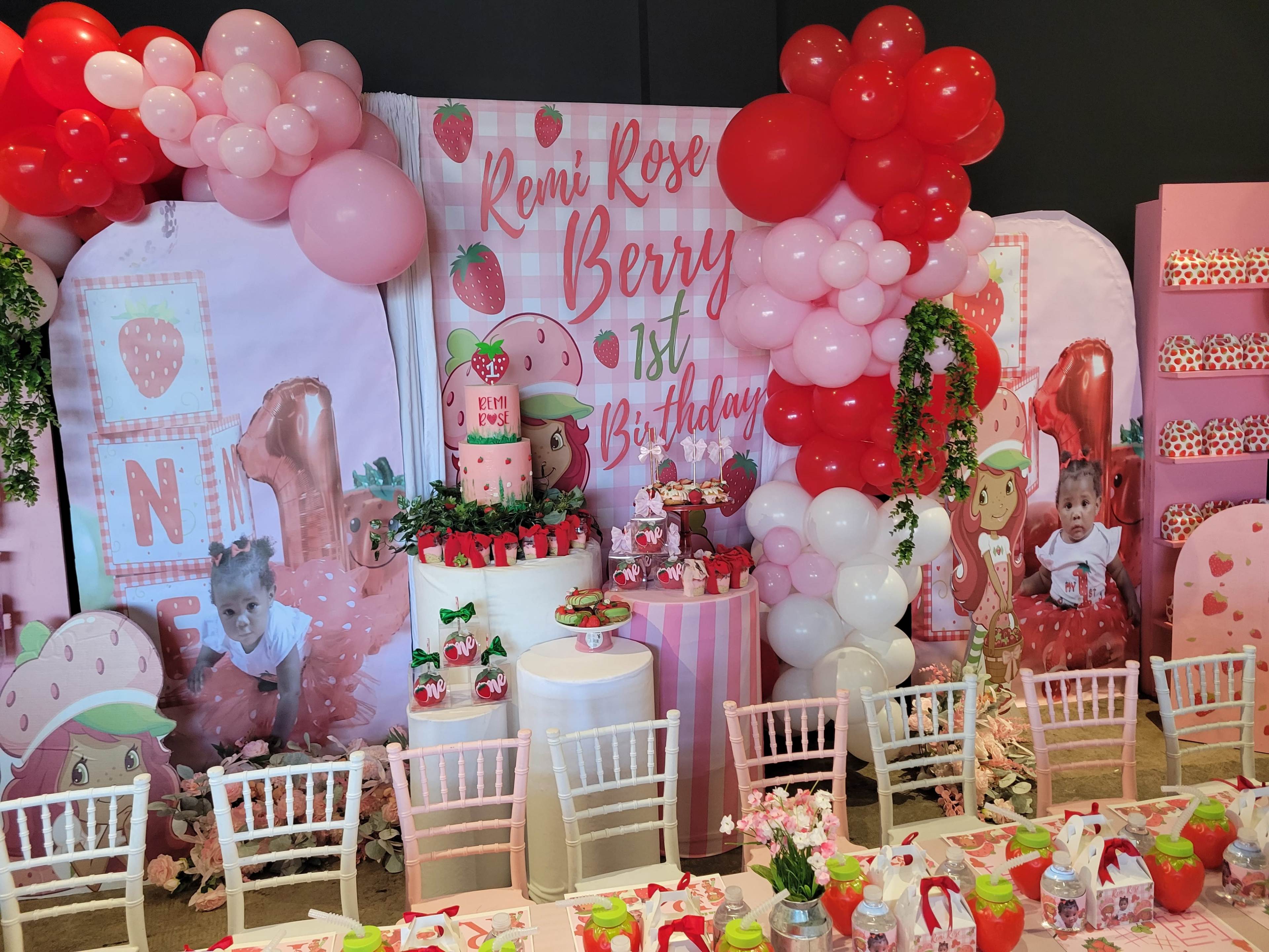 A colorful birthday party setup featuring a decorated backdrop with strawberries and balloons, a cake on a table, and themed decorations surrounding a dining area.