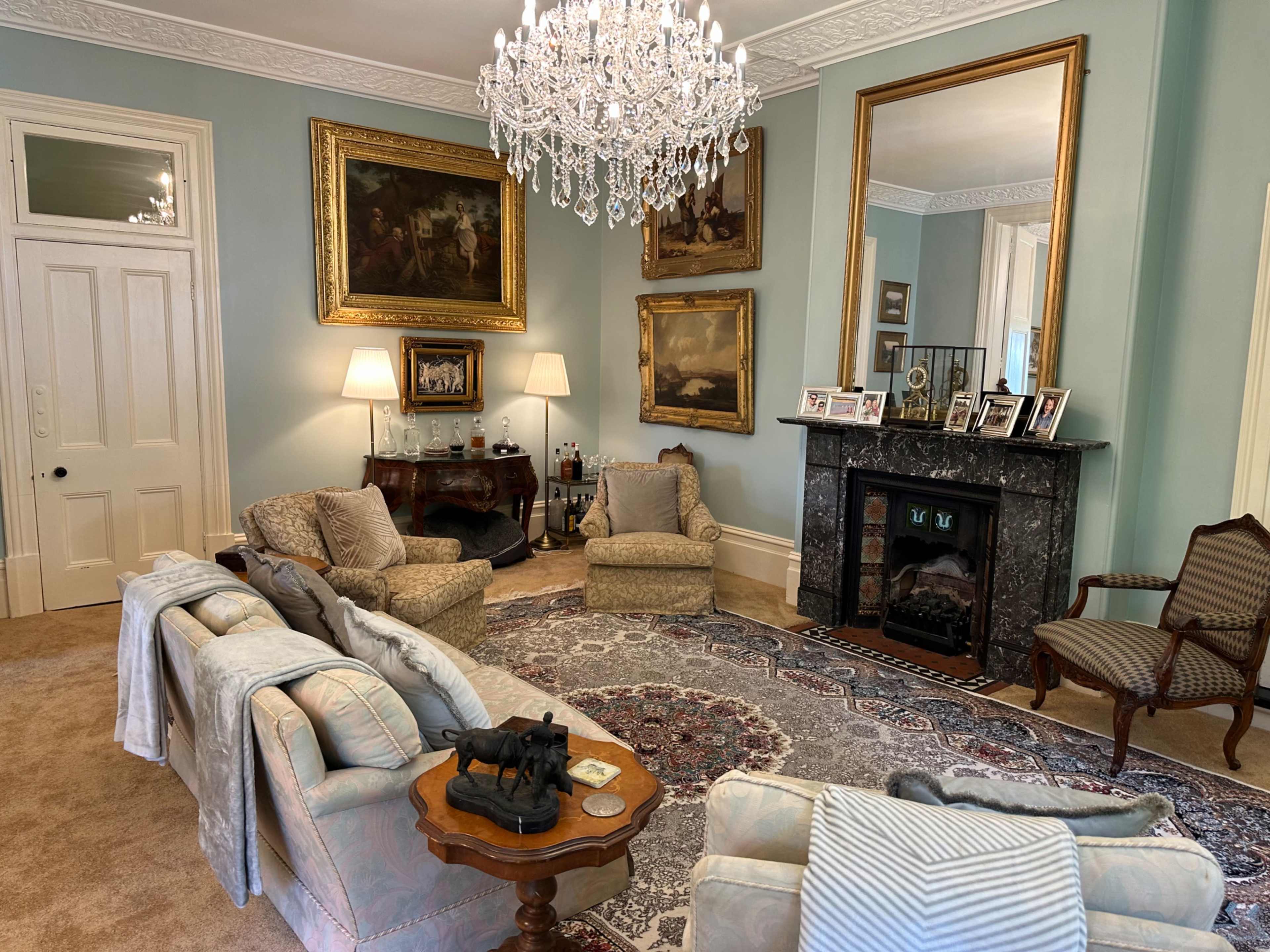 The living room features a large ornate chandelier, elegant seating, a marble fireplace, and framed artwork on the walls.