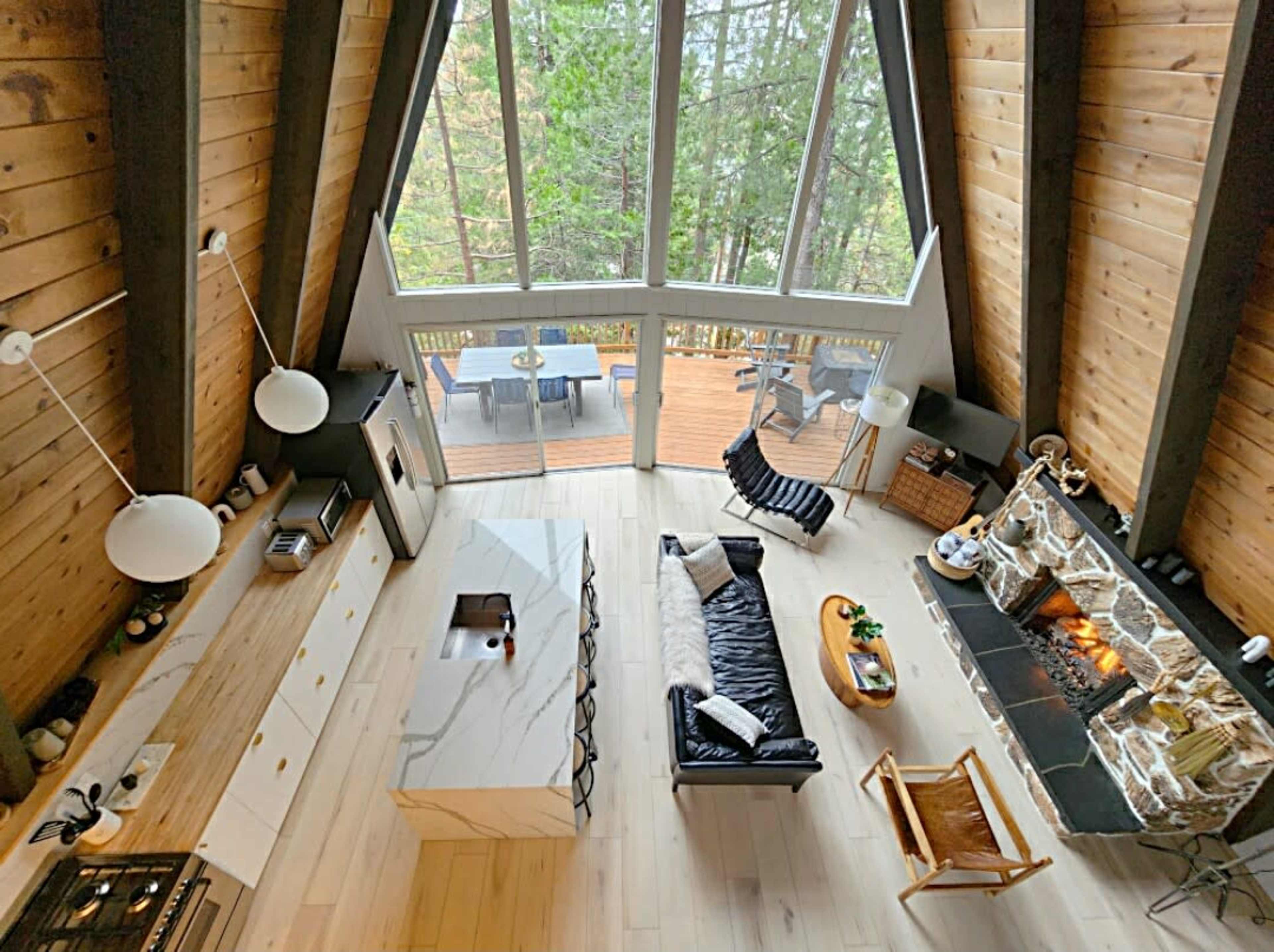 The image shows a modern, A-frame cabin interior with a spacious living area, a large stone fireplace, and large windows overlooking a wooden deck surrounded by trees.