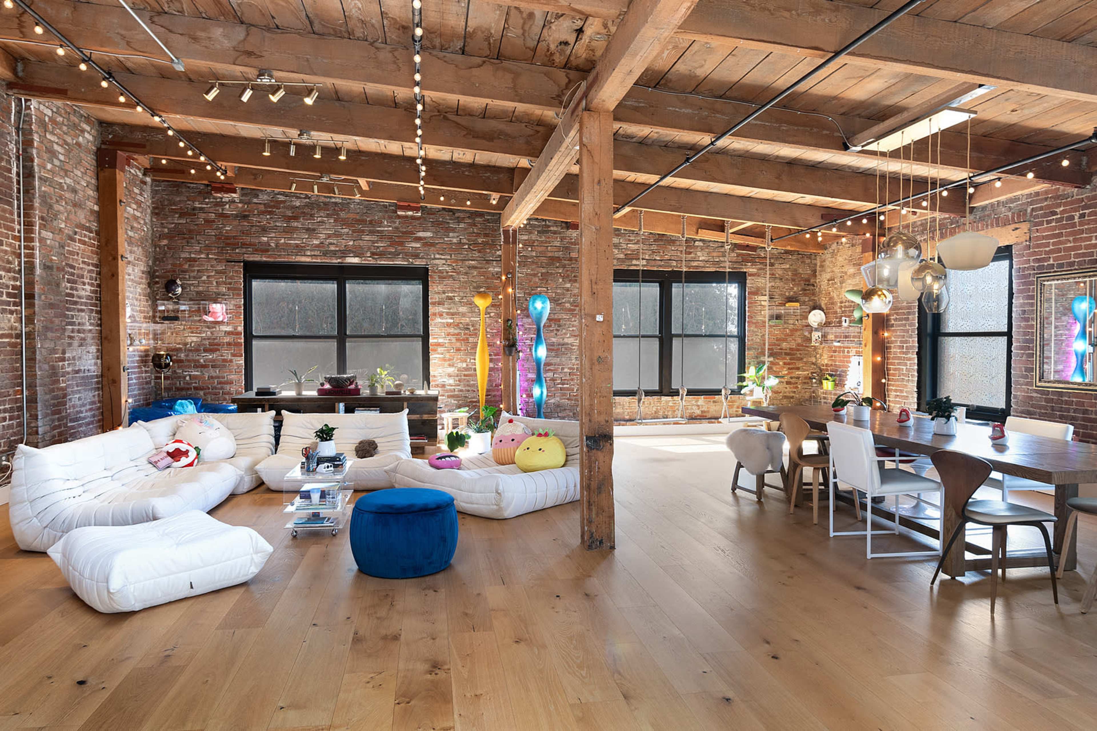 A spacious, modern interior with exposed brick walls, wooden beams, a seating area with white couches, and a dining table surrounded by chairs.