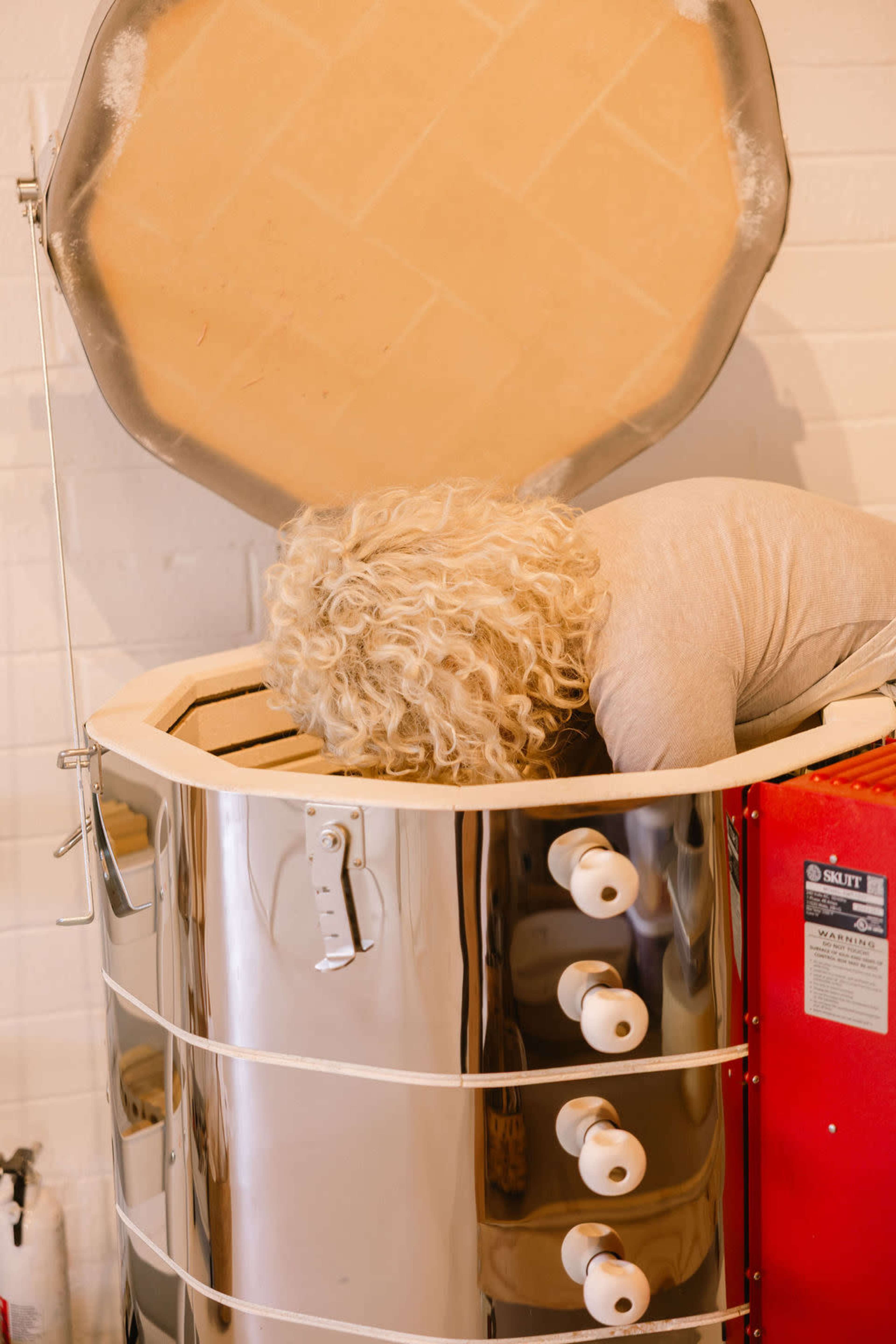 A person with curly hair is leaning into a large, shiny kiln with an open lid.