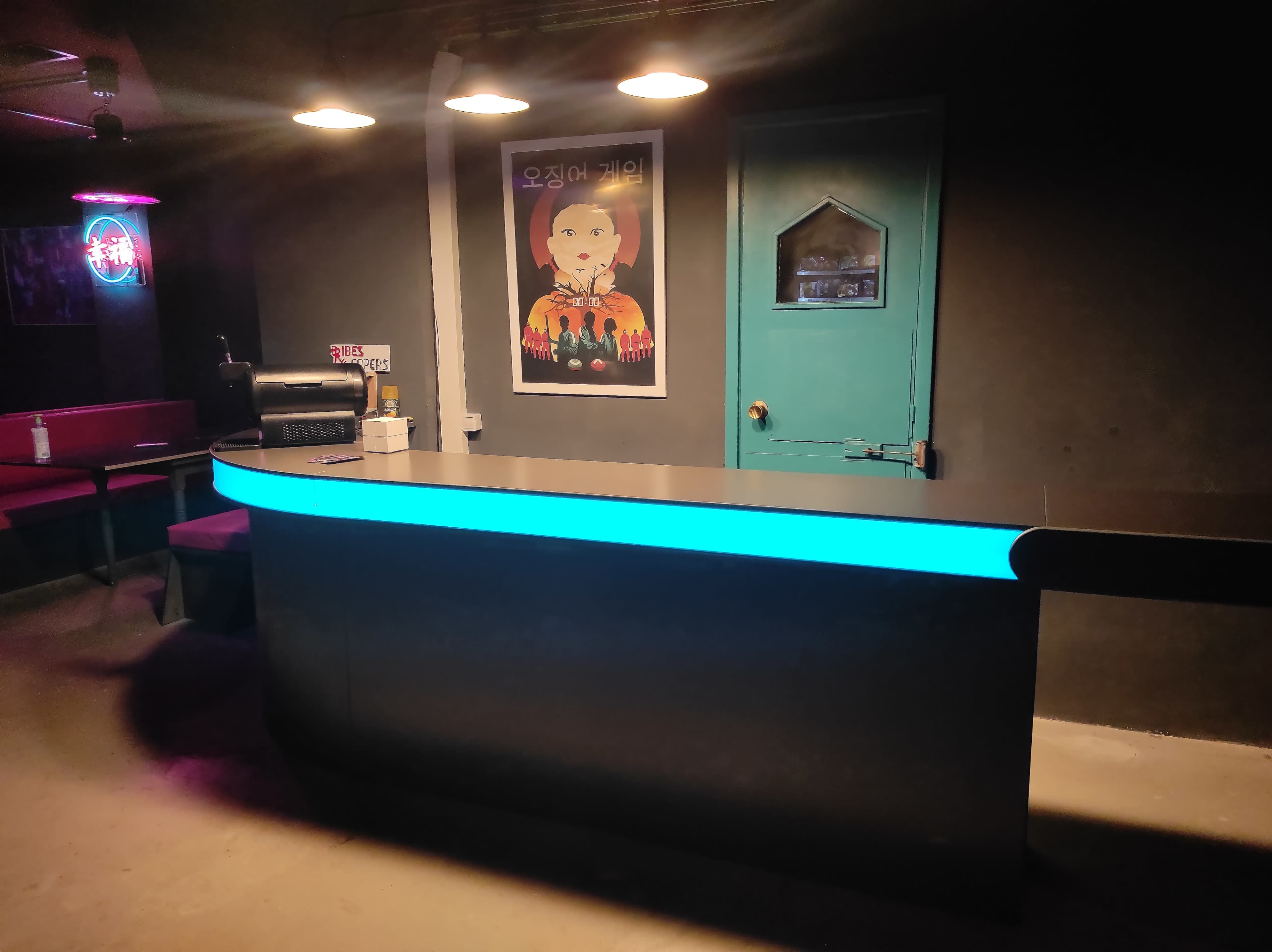 A dimly lit bar area with a curved countertop illuminated by a blue light, featuring a poster and a green door in the background.