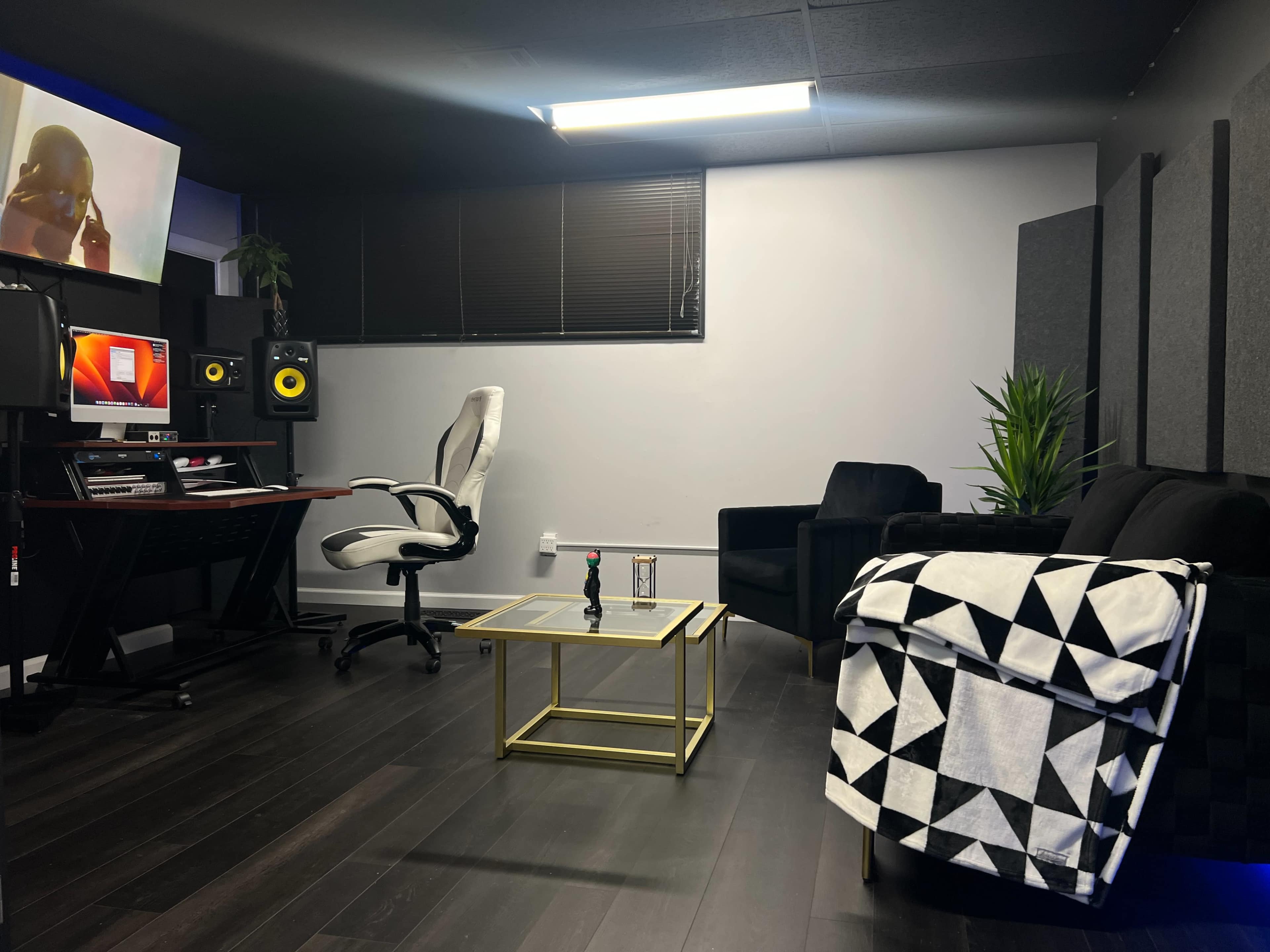 A modern recording studio with a desk, computer, sound monitors, a comfortable chair, and a black couch, all set against dark flooring and gray acoustic panels.
