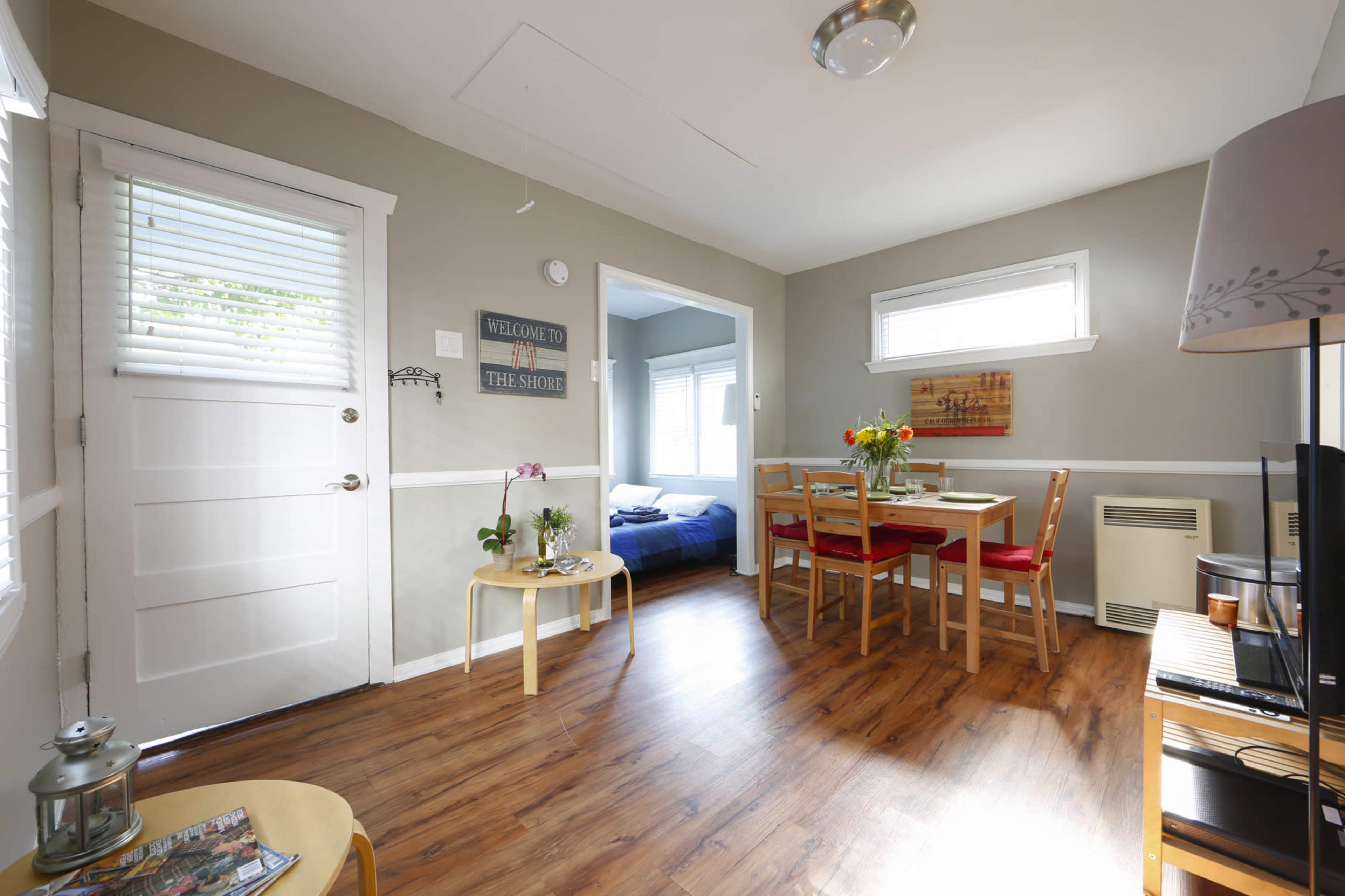 A small, well-lit living area with a dining table, a plant, and a view into a bedroom through an open doorway.