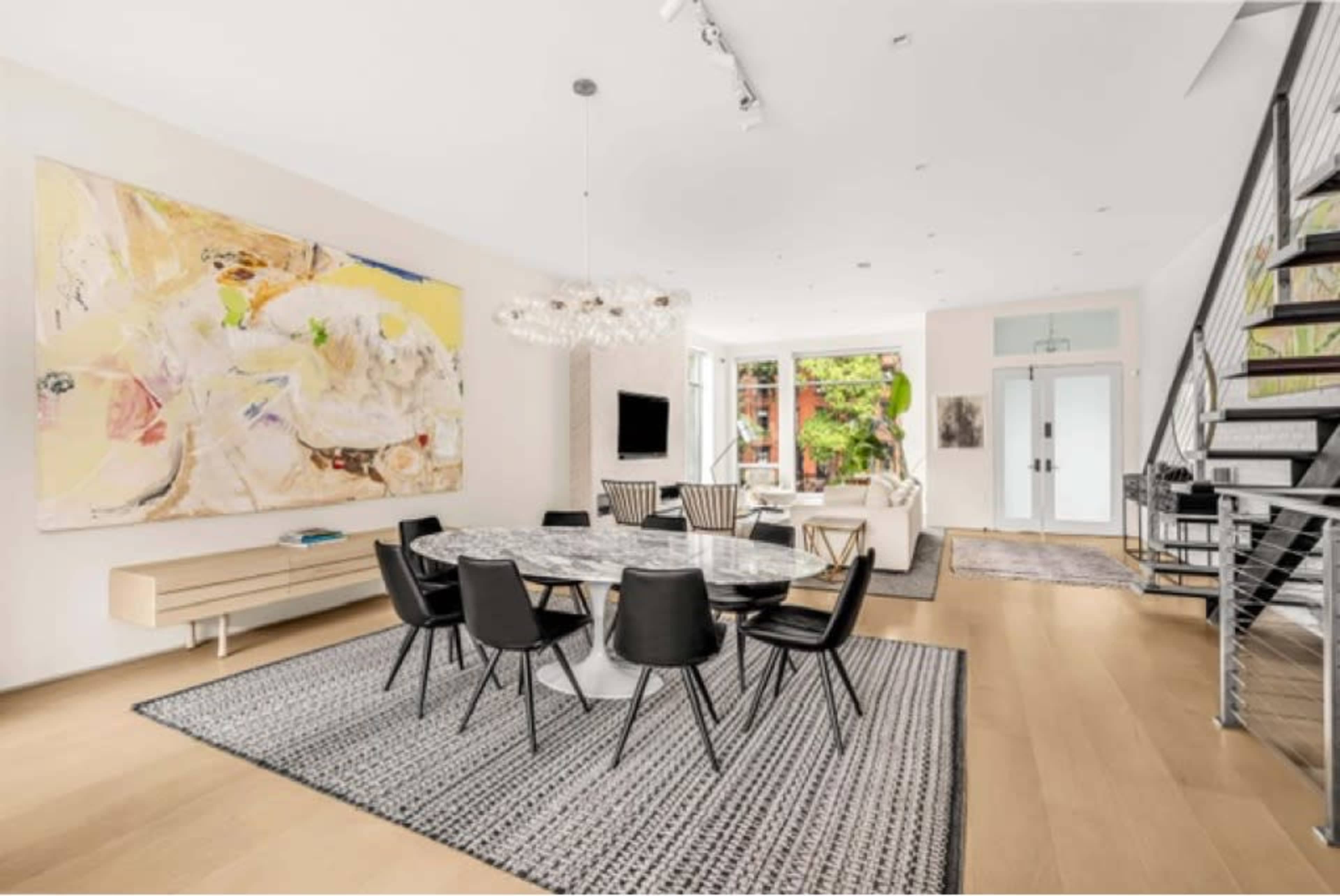 A modern dining area features a round marble table surrounded by black chairs, an abstract painting on the wall, and large windows that provide natural light.