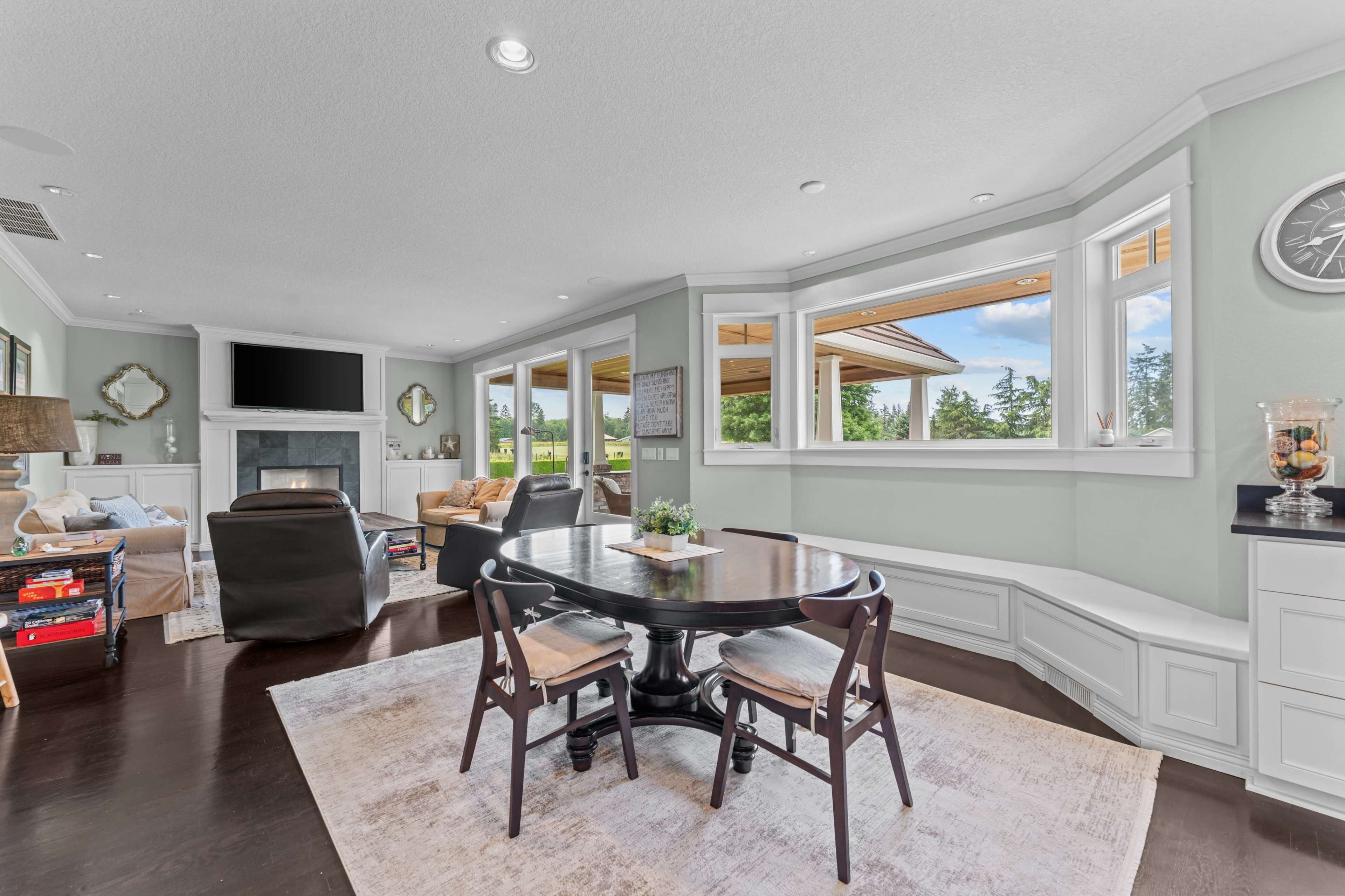 The image shows a modern living room with a round dining table and chairs, a fireplace, and large windows that overlook a scenic view.