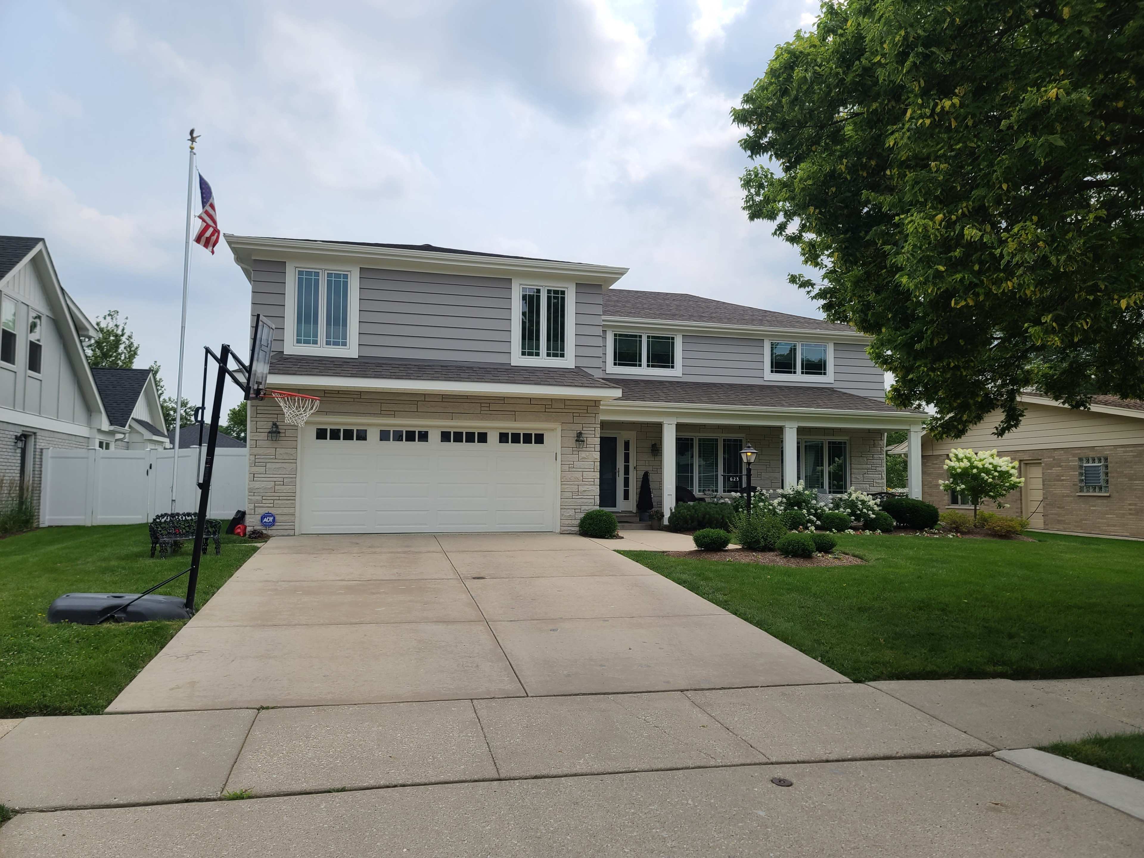 A two-story gray house with a basketball hoop in the driveway and an American flag flying in front.