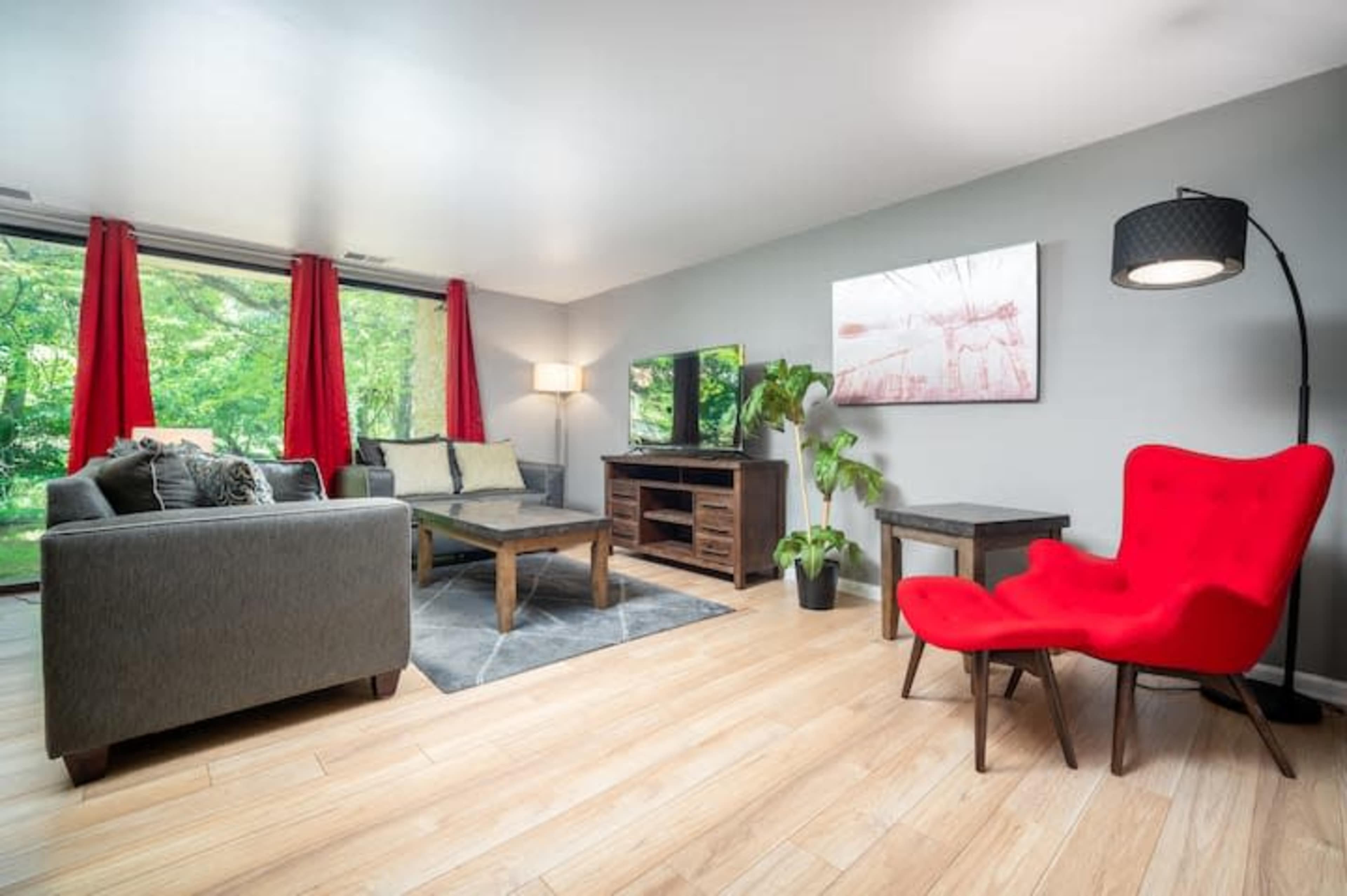 A modern living room with a gray sofa, a red accent chair, a wooden coffee table, and a television, framed by large windows with red curtains overlooking a green outdoor area.