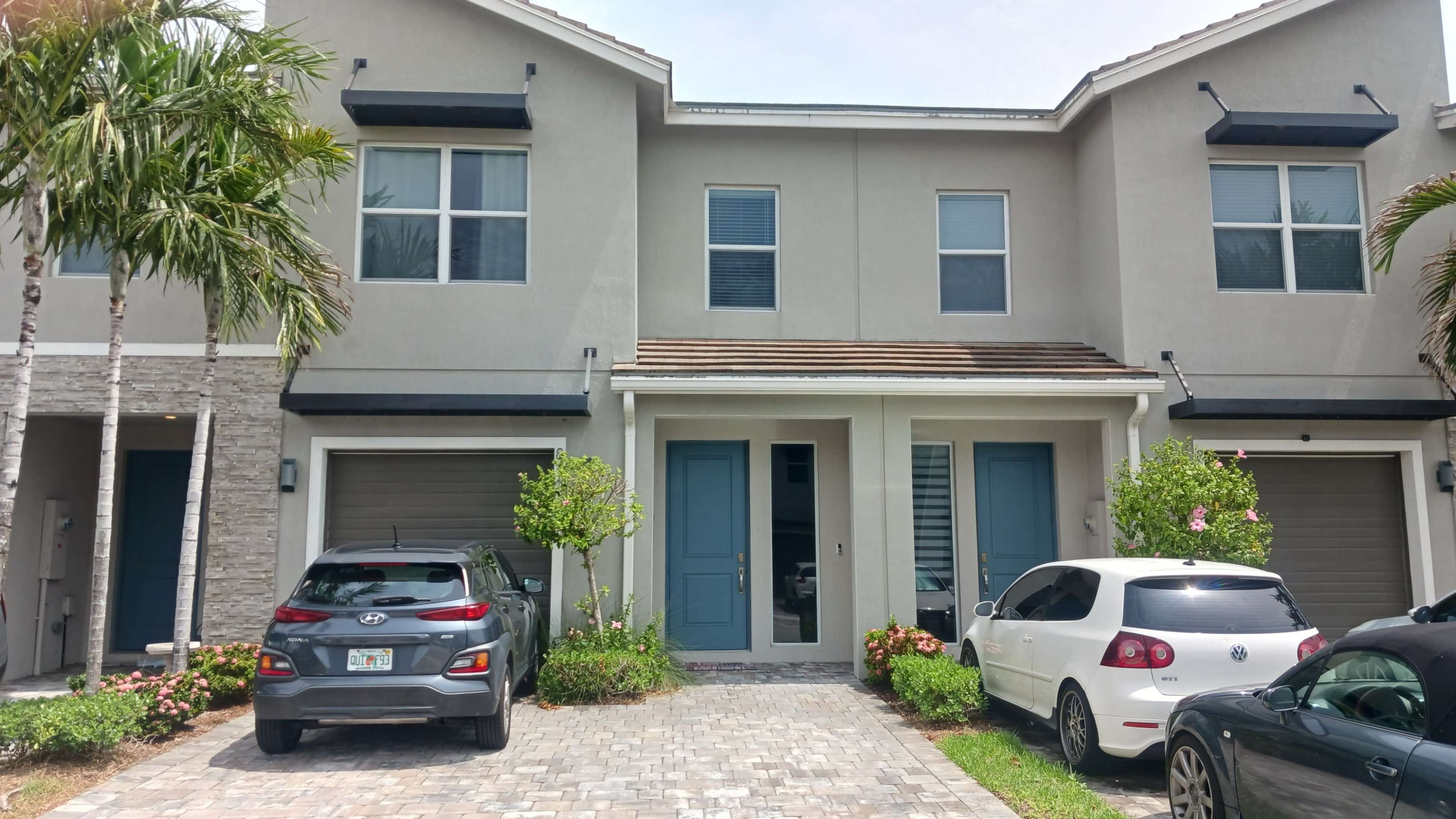 The image shows a multi-unit residential building with two parked cars in front, surrounded by landscaping and palm trees.