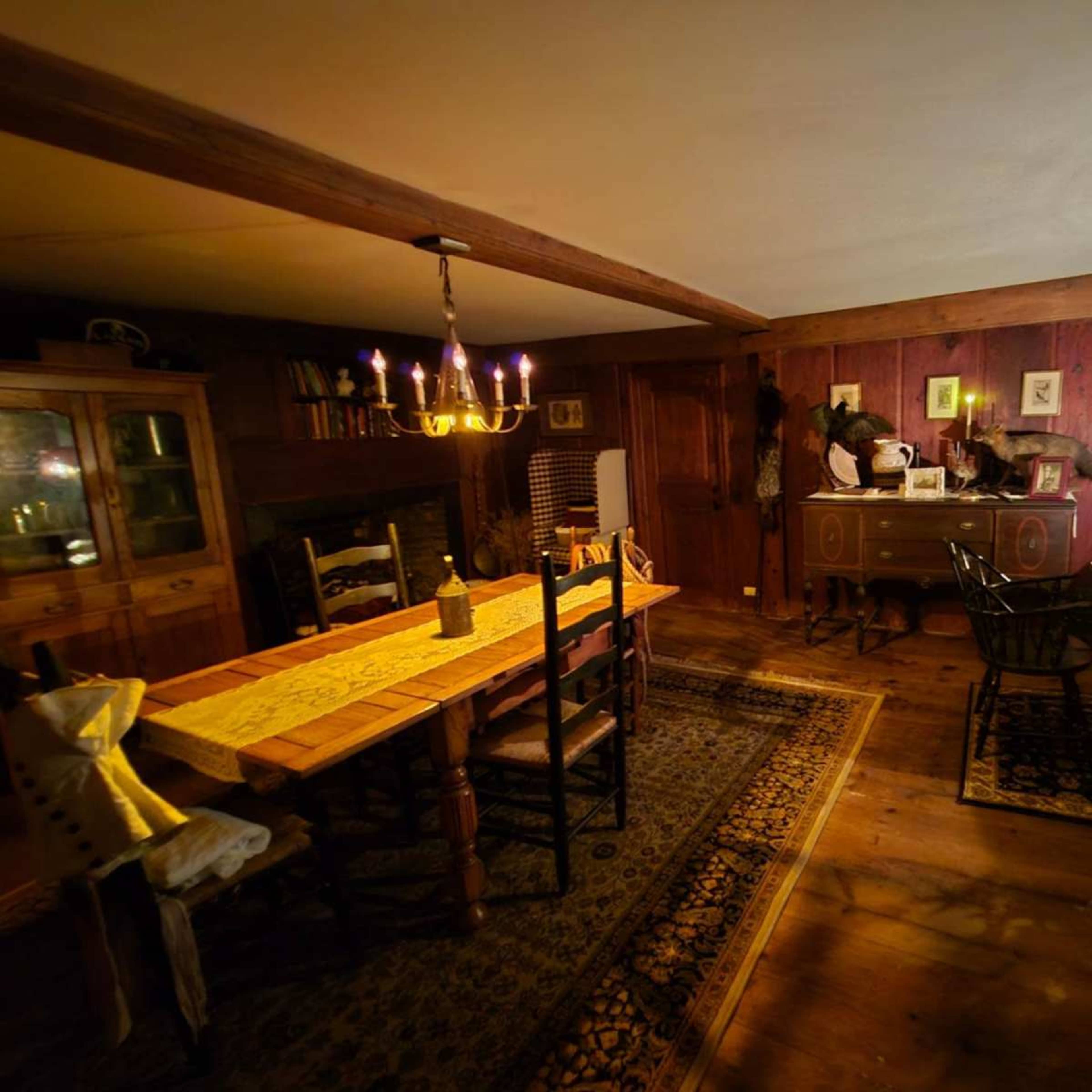 The image depicts a rustic dining room featuring a long wooden table with chairs, a chandelier, and wooden walls adorned with framed pictures and decor.