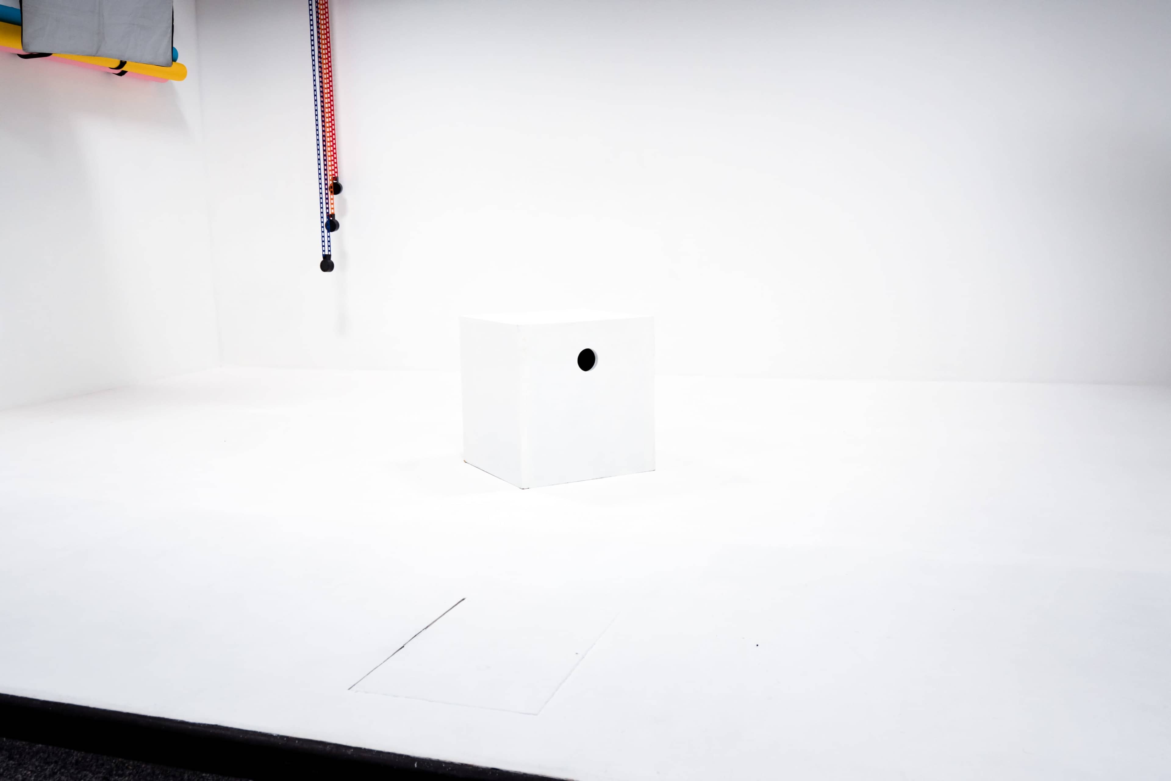 A white box with a circular hole is placed in the center of a clean, empty white room.