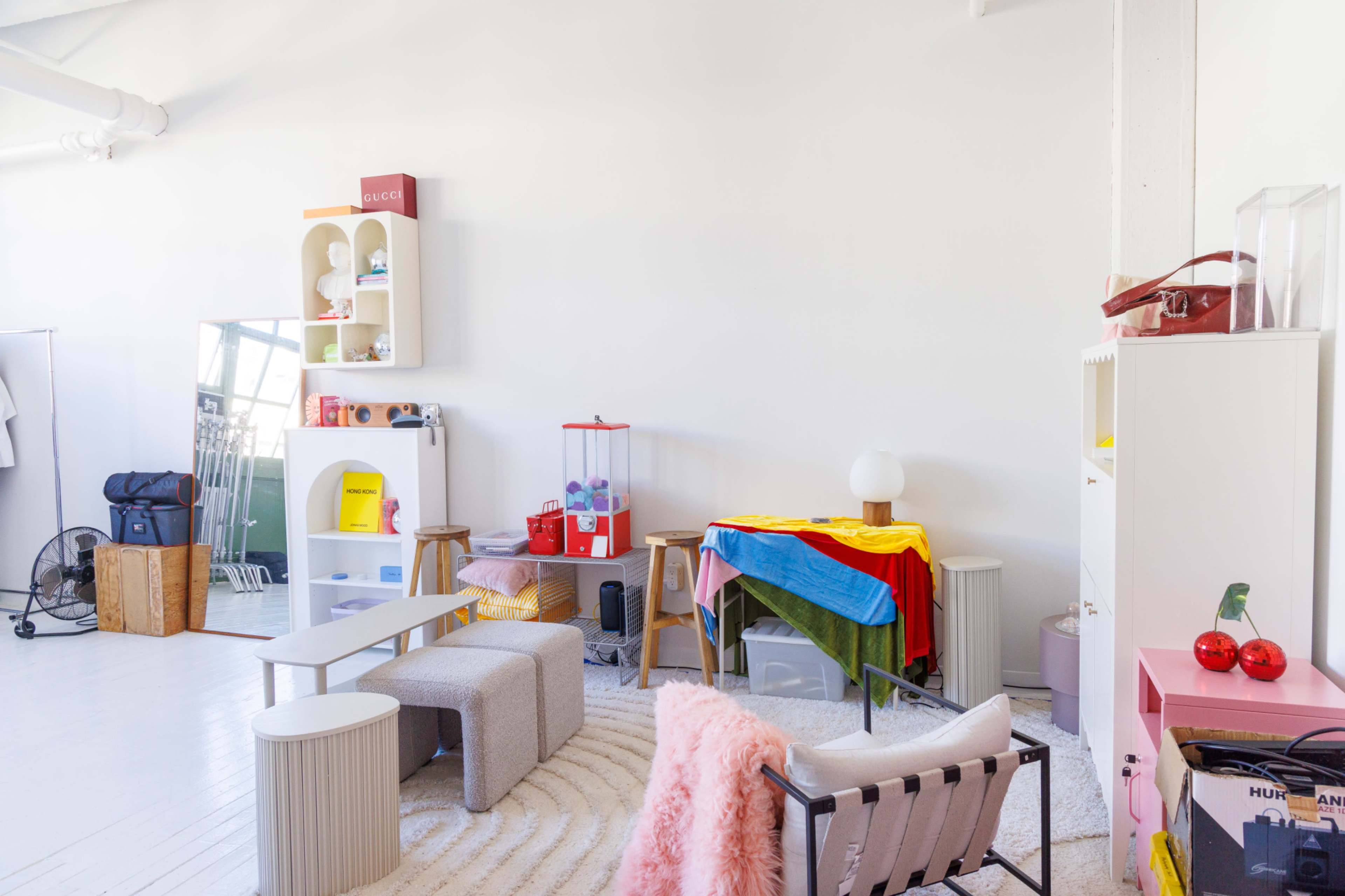 The image shows a brightly lit room featuring various furniture pieces, colorful textiles, and decorative items arranged in a modern style.