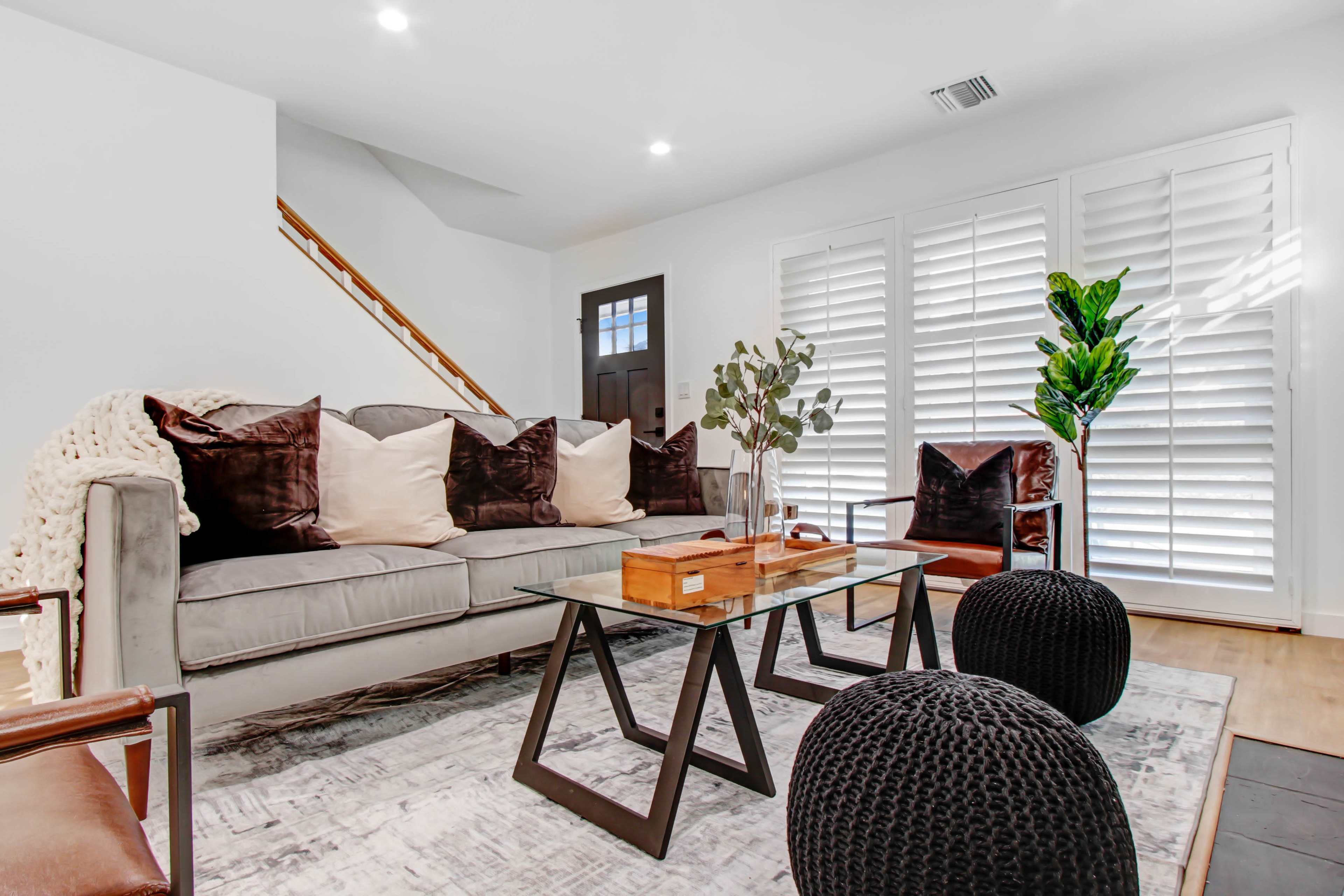 A modern living room features a gray sofa with decorative pillows, a glass coffee table, two black poufs, and potted plants, with plantation shutters on the windows and a staircase in the background.