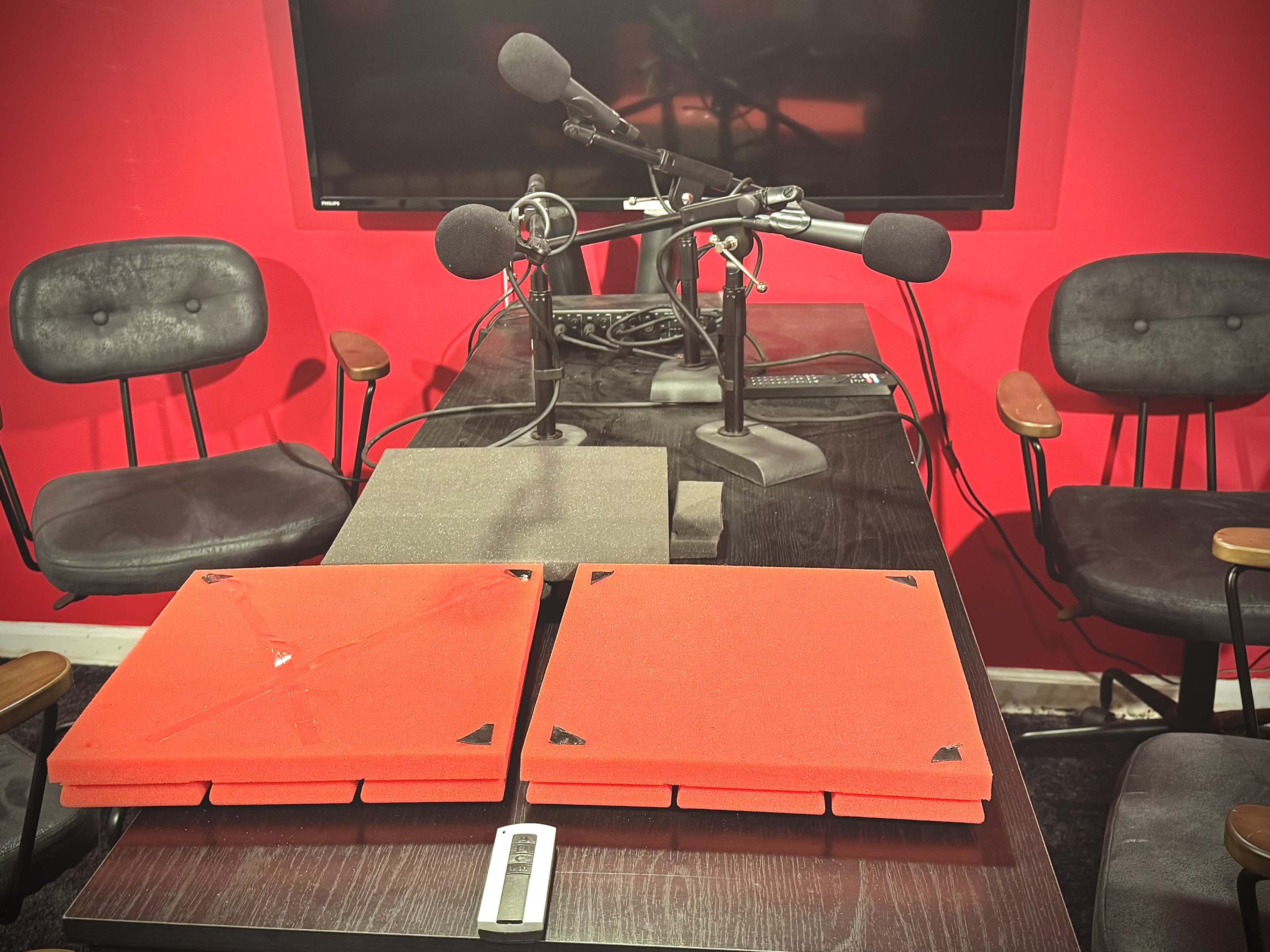 The image shows a recording studio setup with two microphone stands, four chairs, and two red soundproofing panels on a black table.