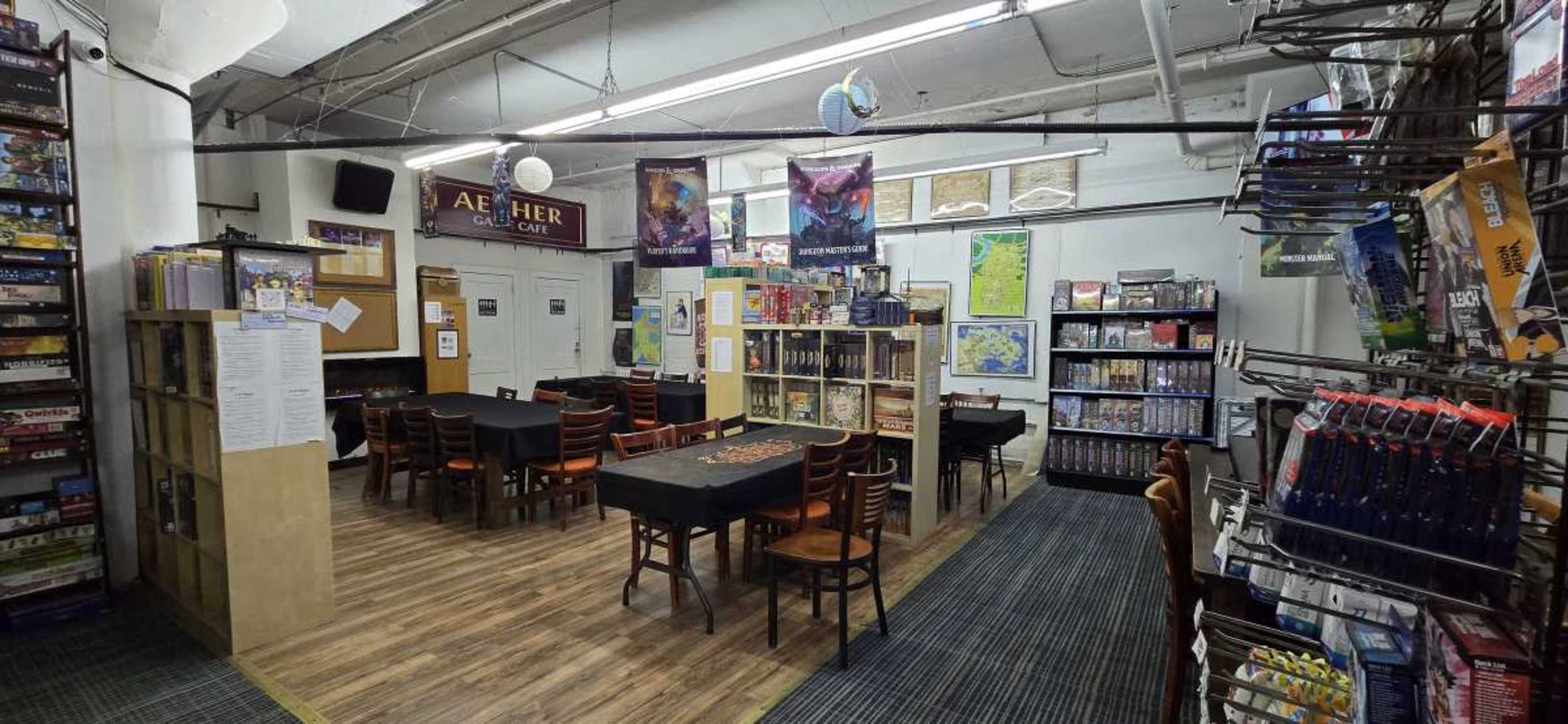 The image shows a board game café with tables arranged for play, shelves stocked with games, and a cozy seating area.