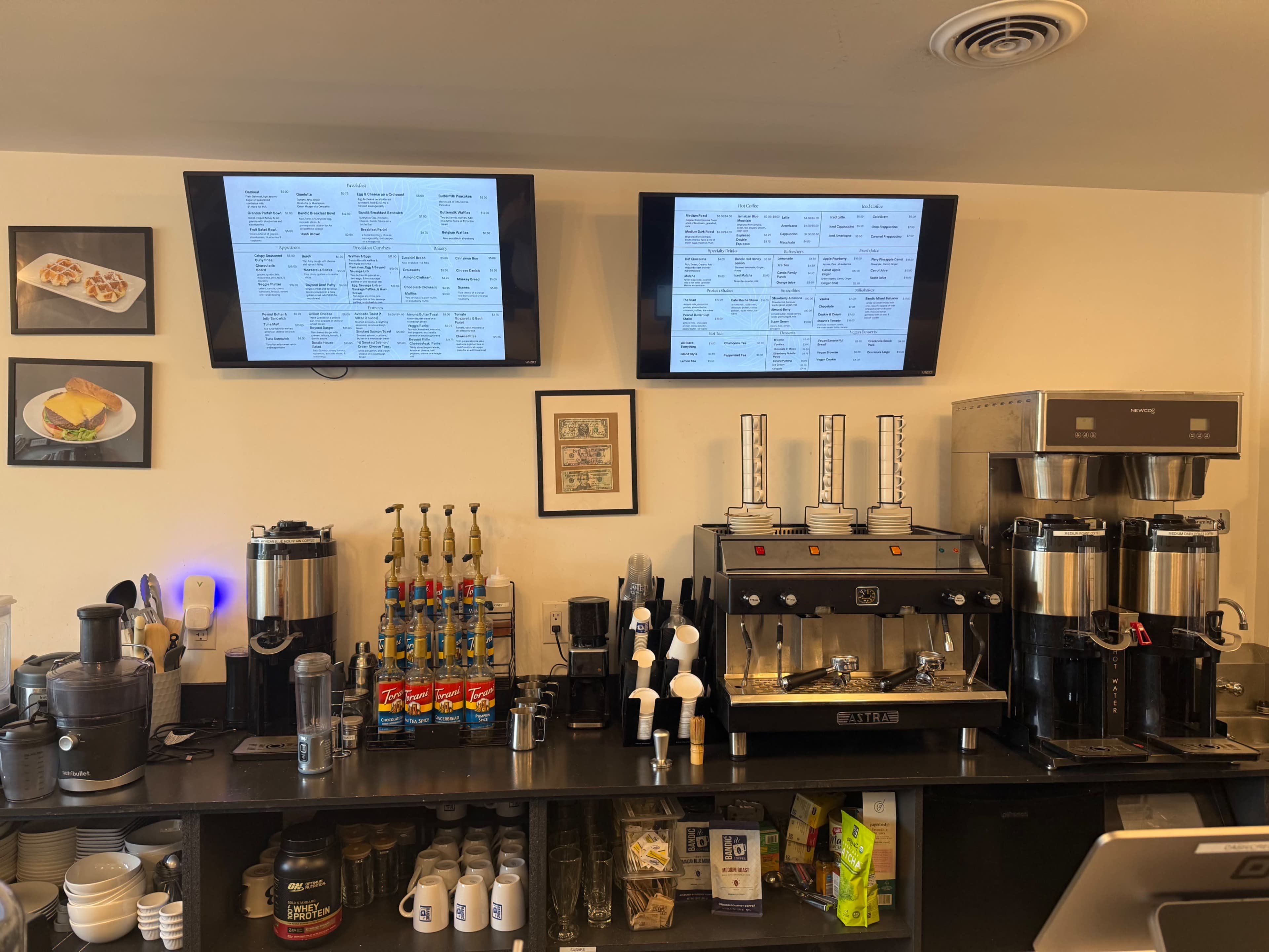 The image shows a coffee shop counter with two digital menu screens, a coffee machine, beverage dispensers, and various ingredients lined up on the shelf.