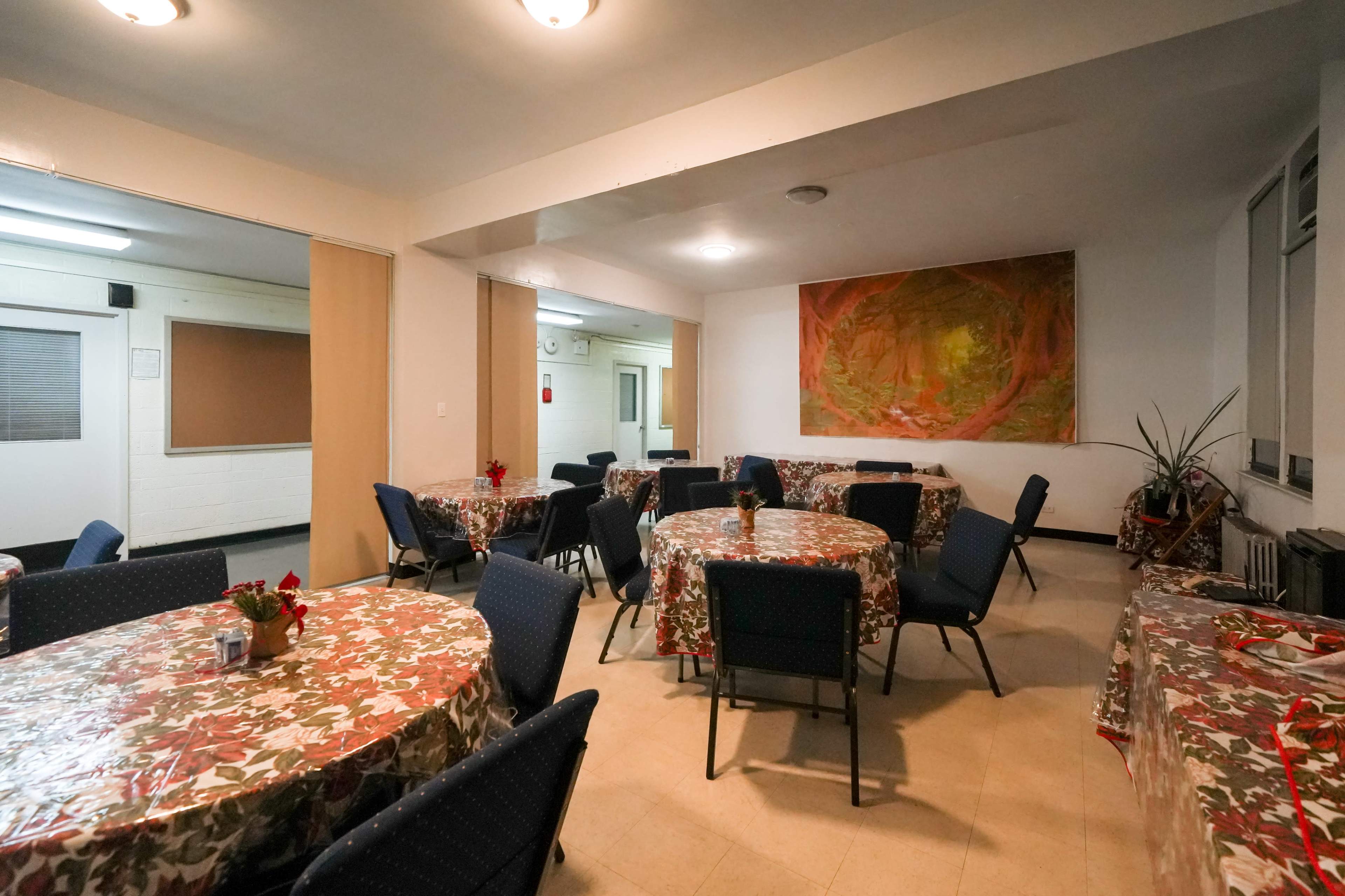 The interior of a community room features several round tables covered with floral tablecloths and chairs arranged around them, along with a large mural on one wall.
