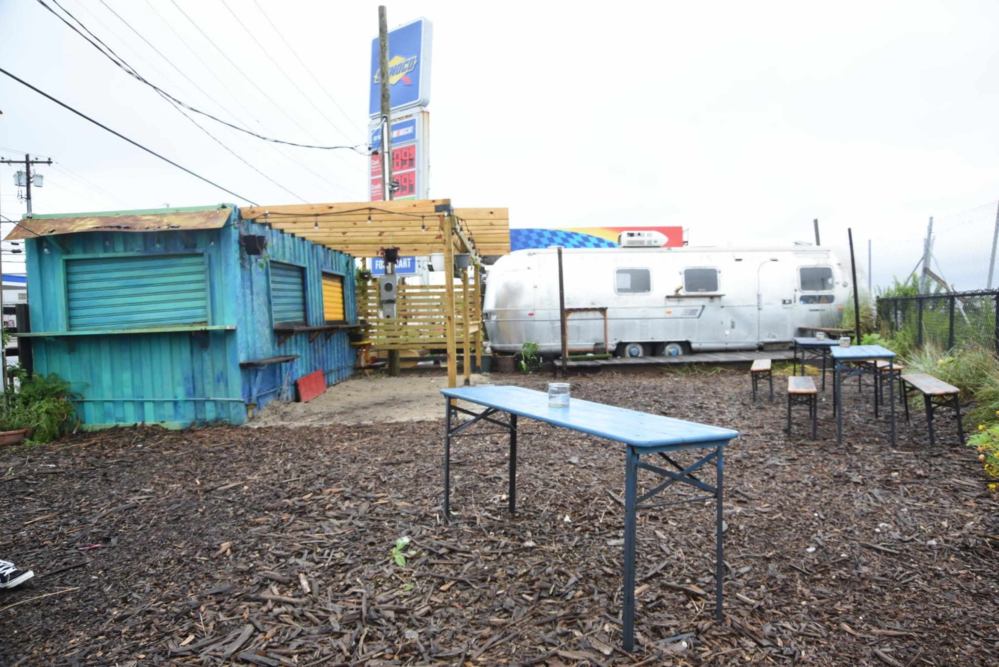 A colorful wooden structure and an Airstream trailer in an outdoor area with several metal tables and a gravel ground.