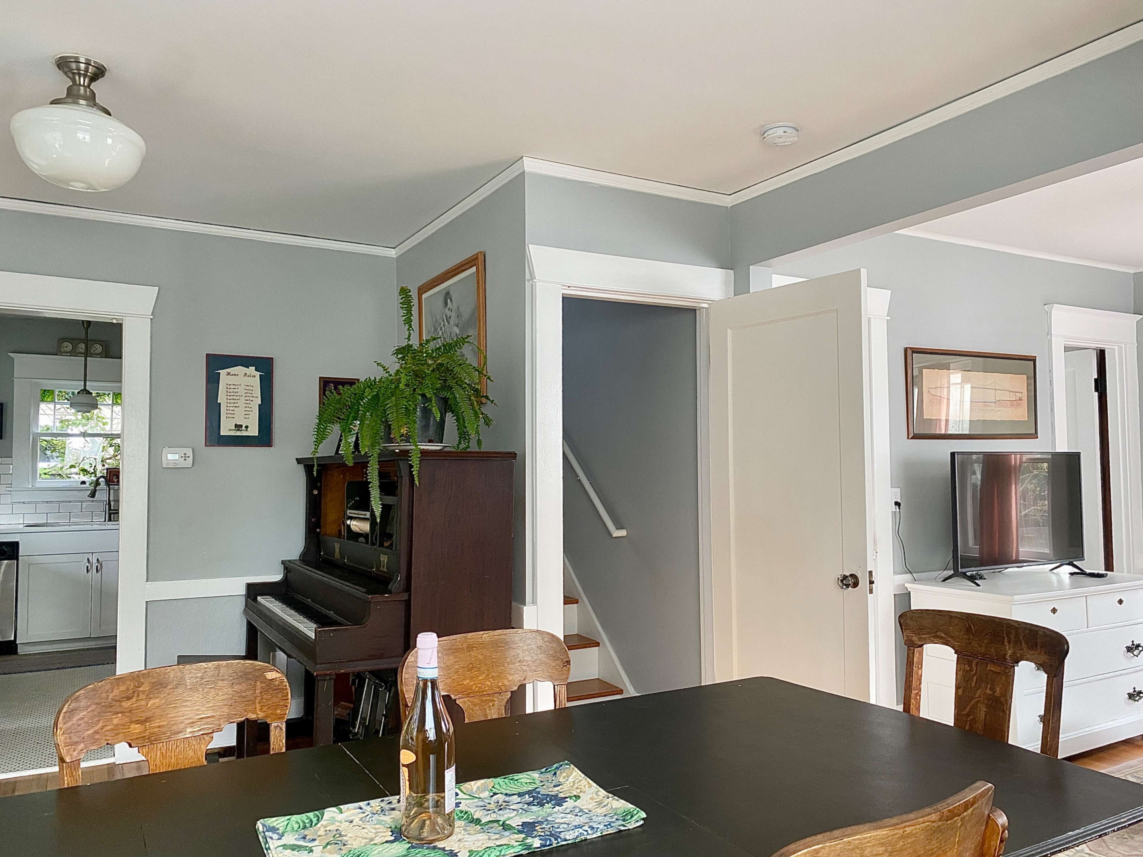 A dining area features a dark wooden table, a bottle of wine, and a piano near a staircase, with gray walls and a window leading to the kitchen.