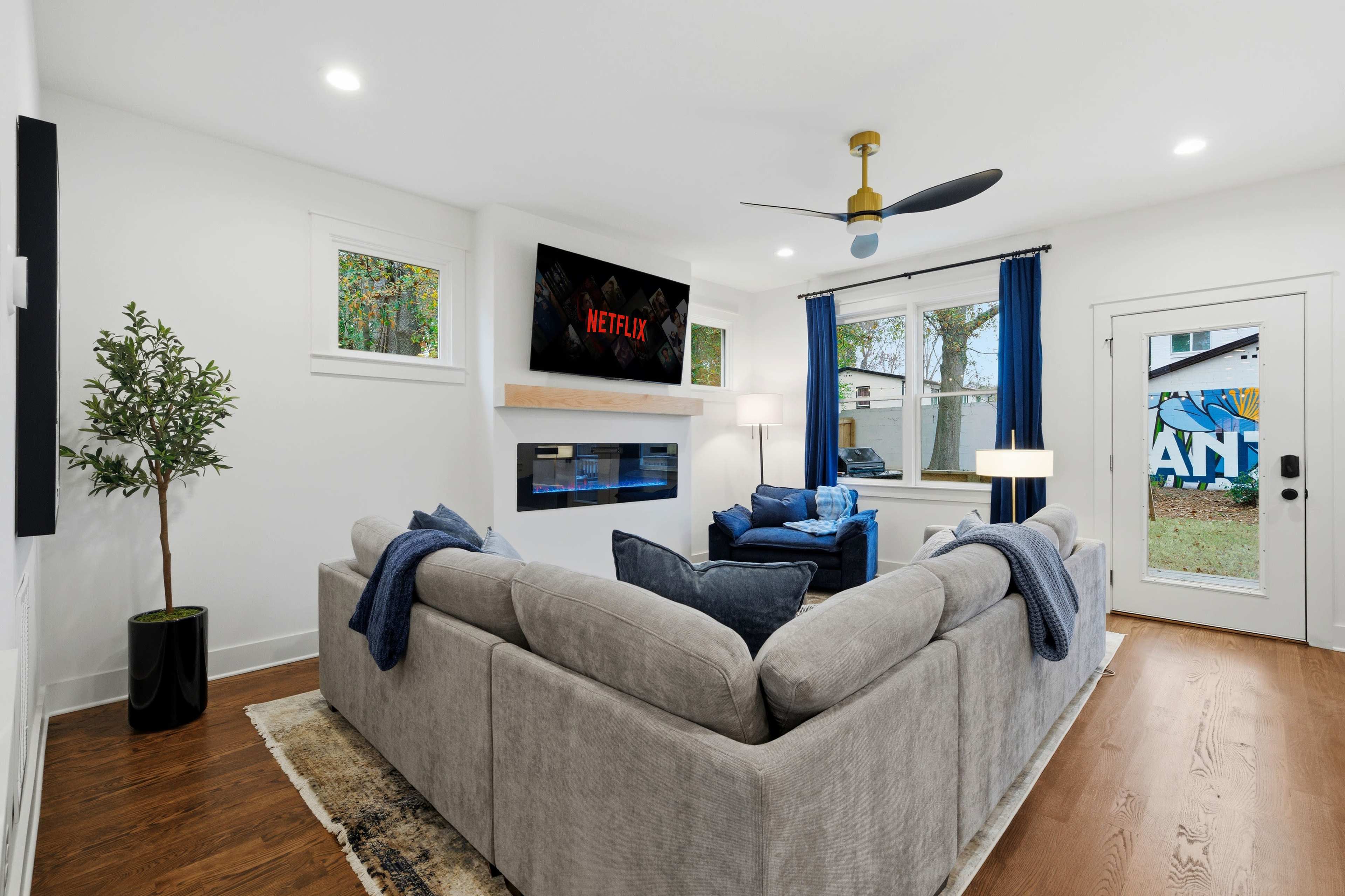 A modern living room features a gray sectional sofa, a mounted TV displaying the Netflix logo, blue accent chairs, and large windows overlooking a backyard.