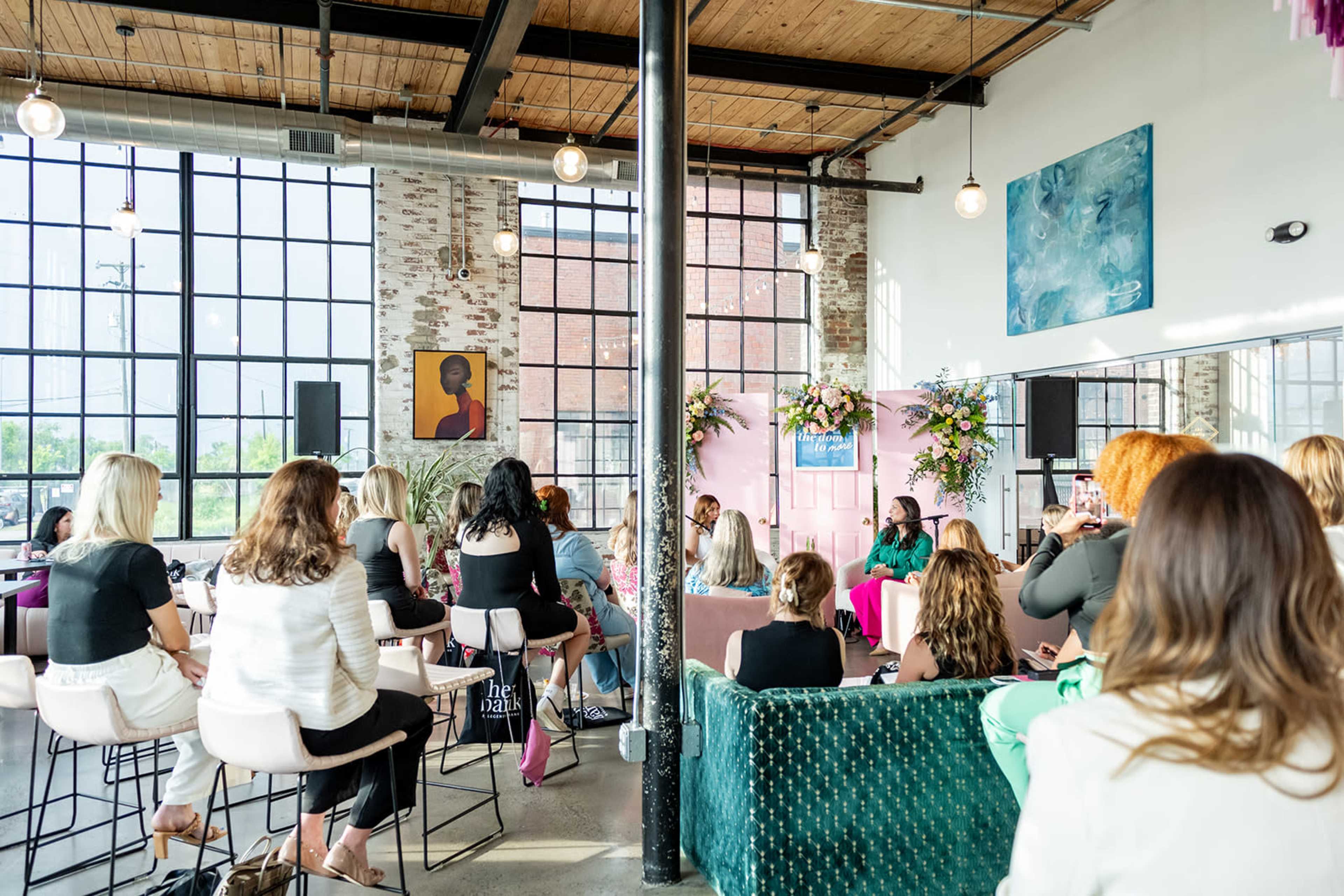 A group of people sits in a modern venue with large windows, attending an event featuring a speaker and floral decorations.