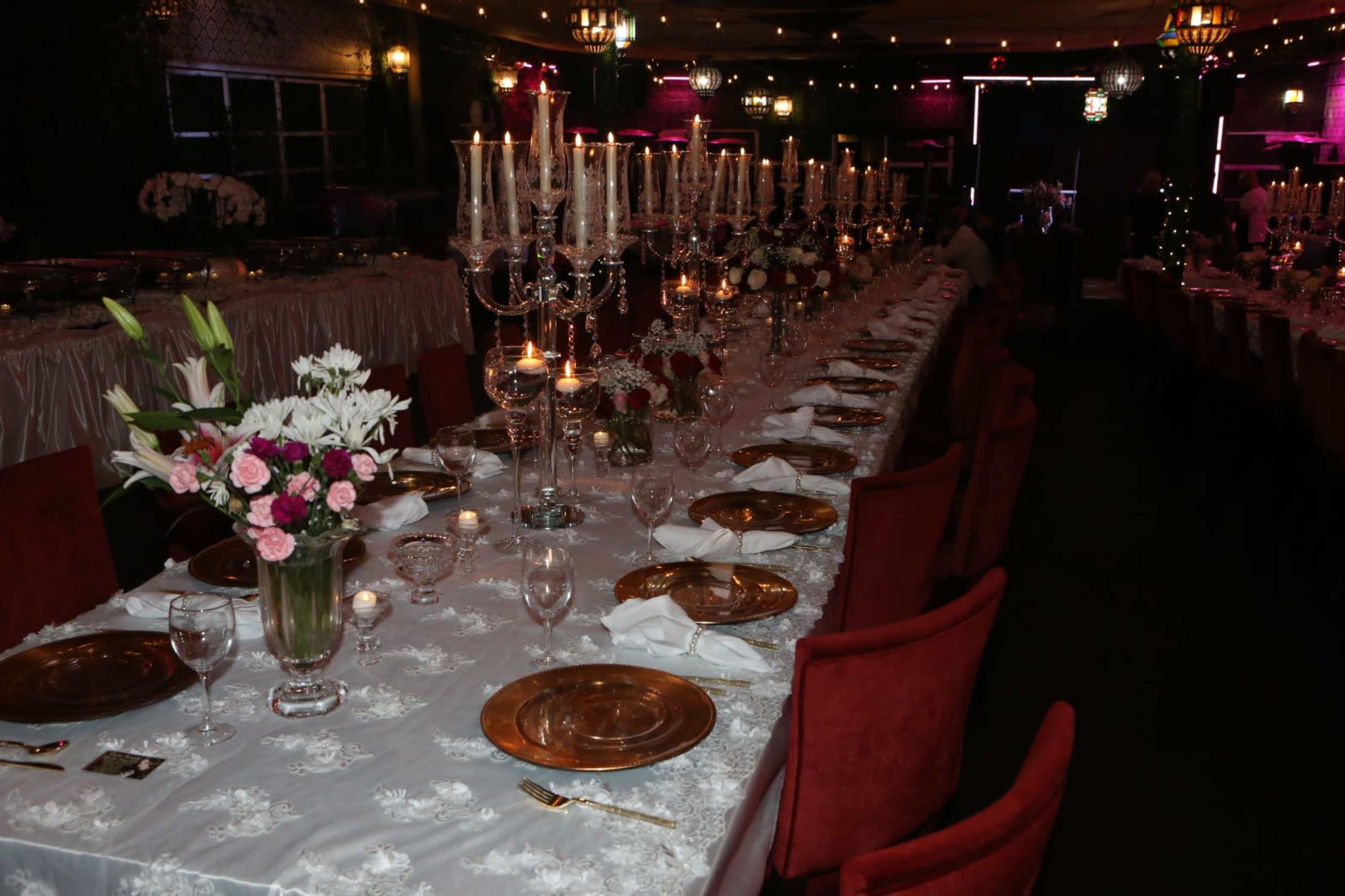 A long dining table elegantly set with golden chargers, white tablecloths, floral arrangements, and multiple candles under soft lighting in a decorated venue.