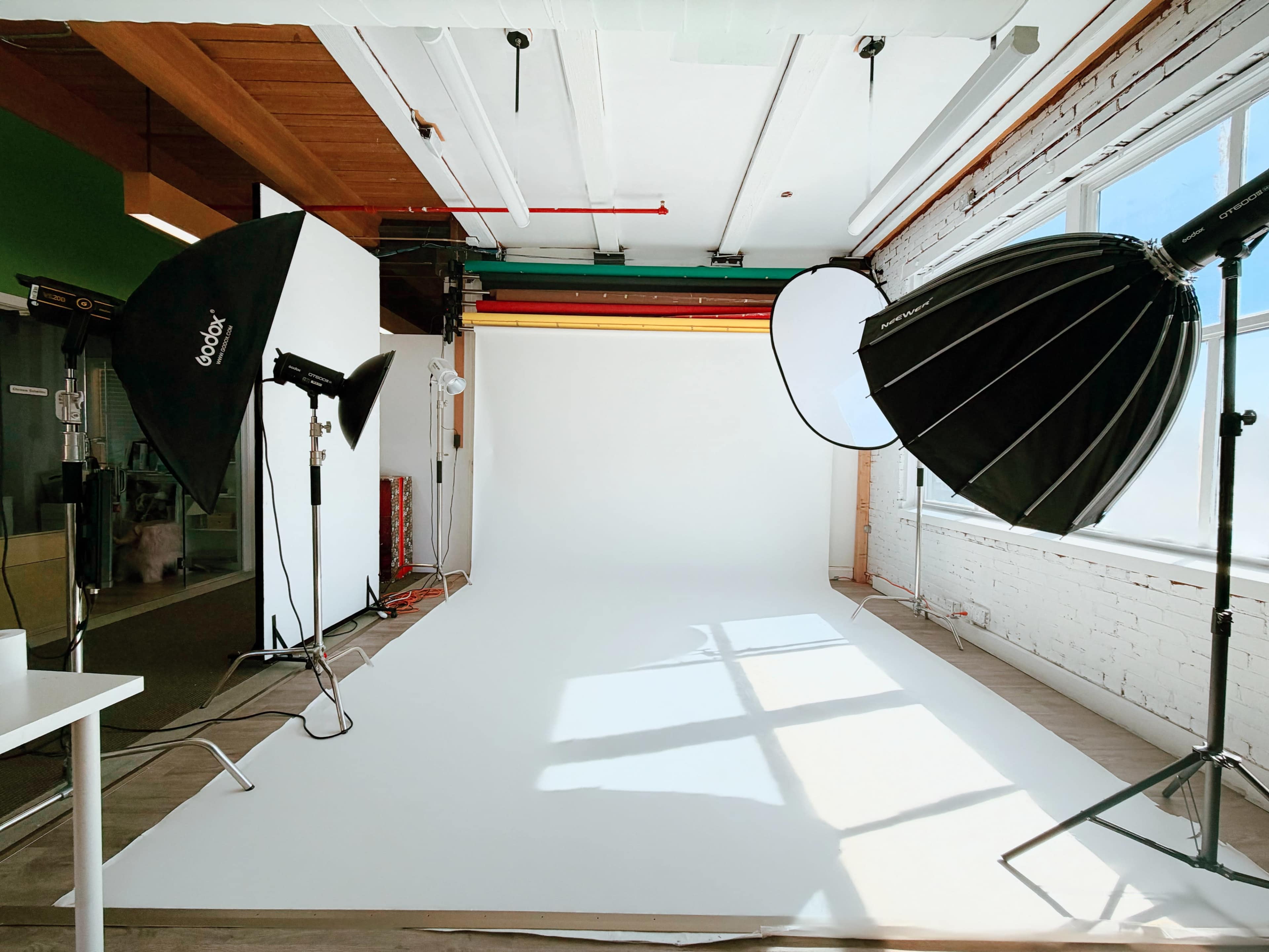 The image shows a photography studio equipped with various lighting setups, a white backdrop, and large windows allowing natural light to enter.
