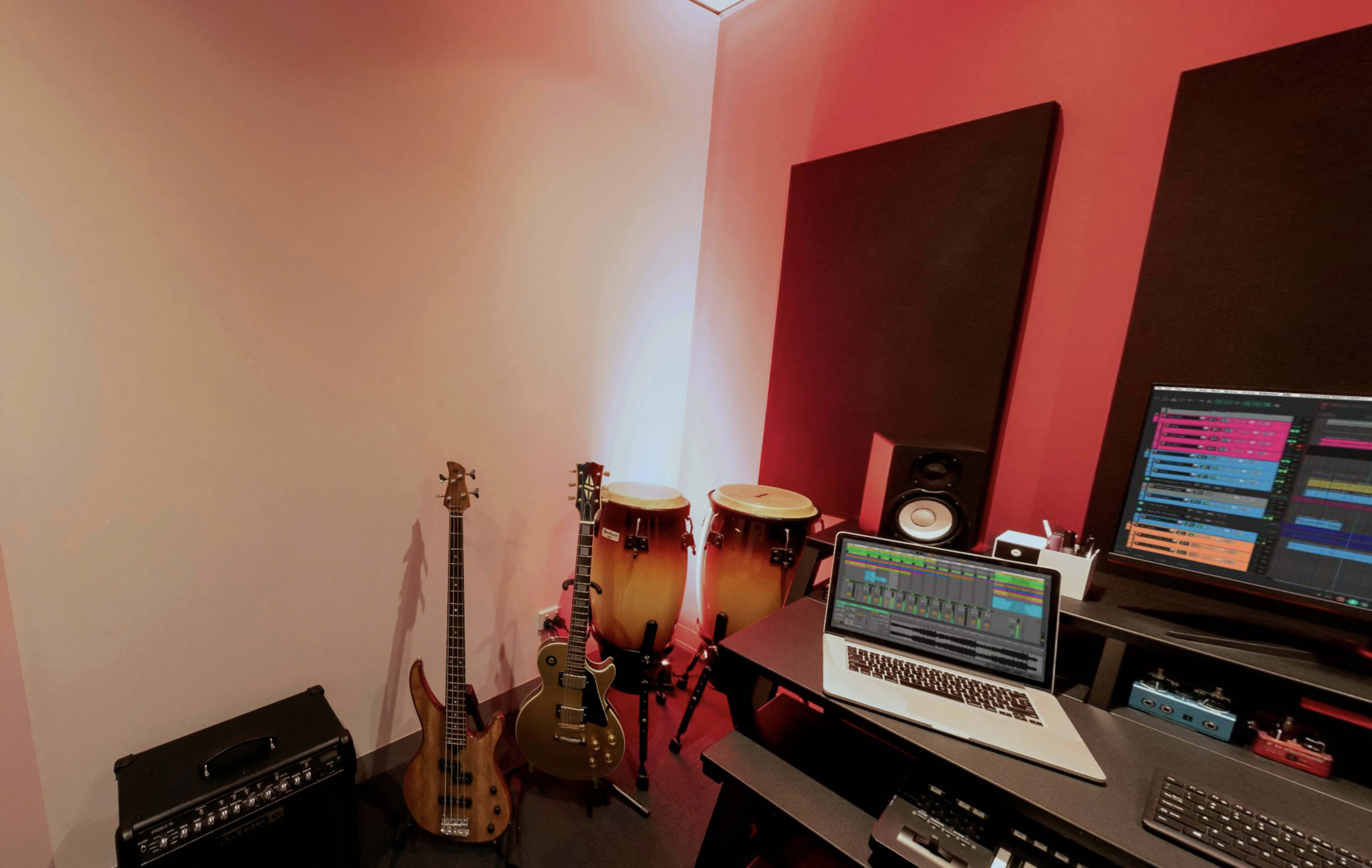A music recording studio with a laptop on a desk, two congas, an electric bass guitar, and an amplifier against a red and white wall.