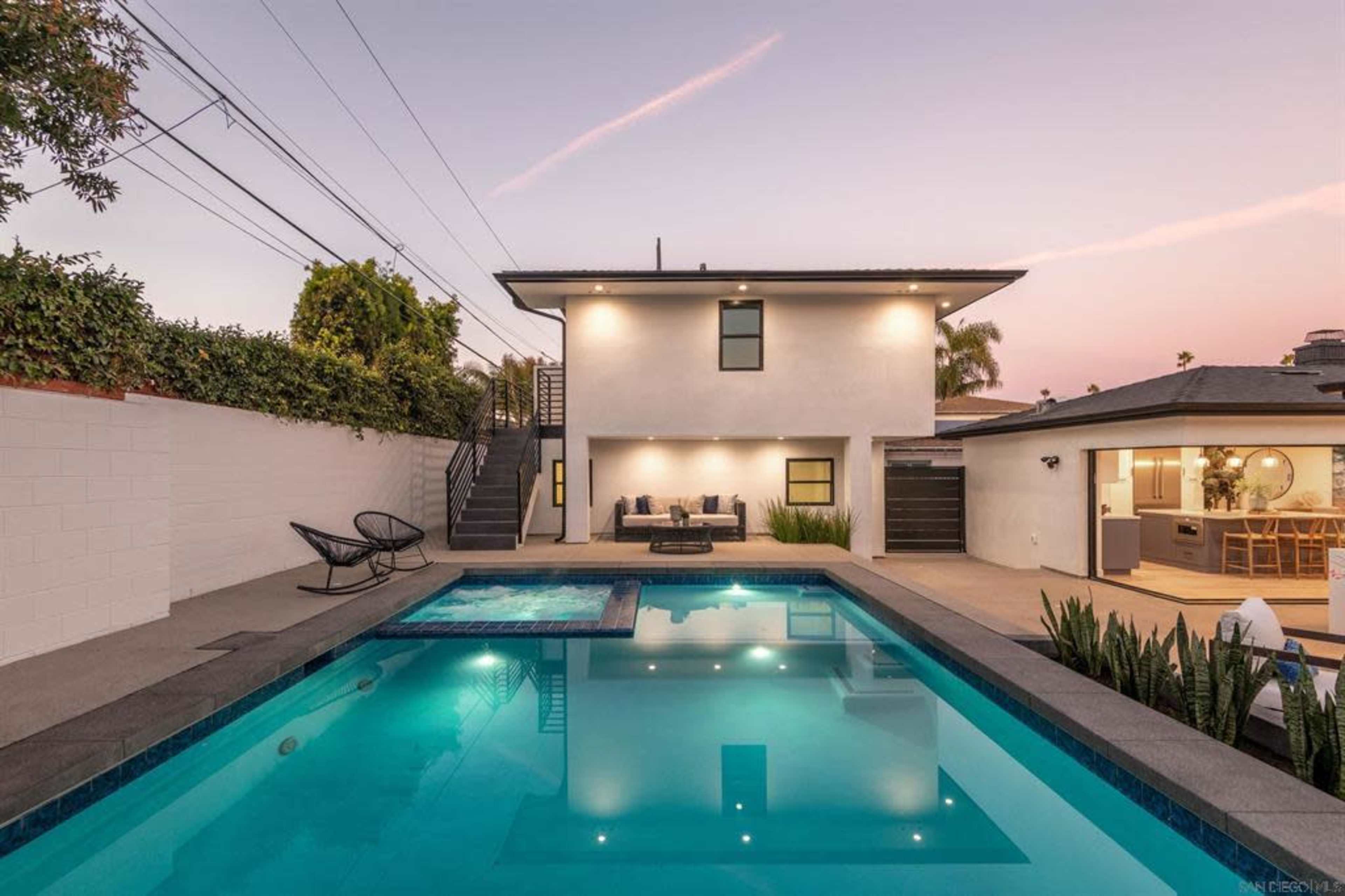 A modern two-story house adjacent to a swimming pool with a hot tub, surrounded by a landscaped yard at dusk.