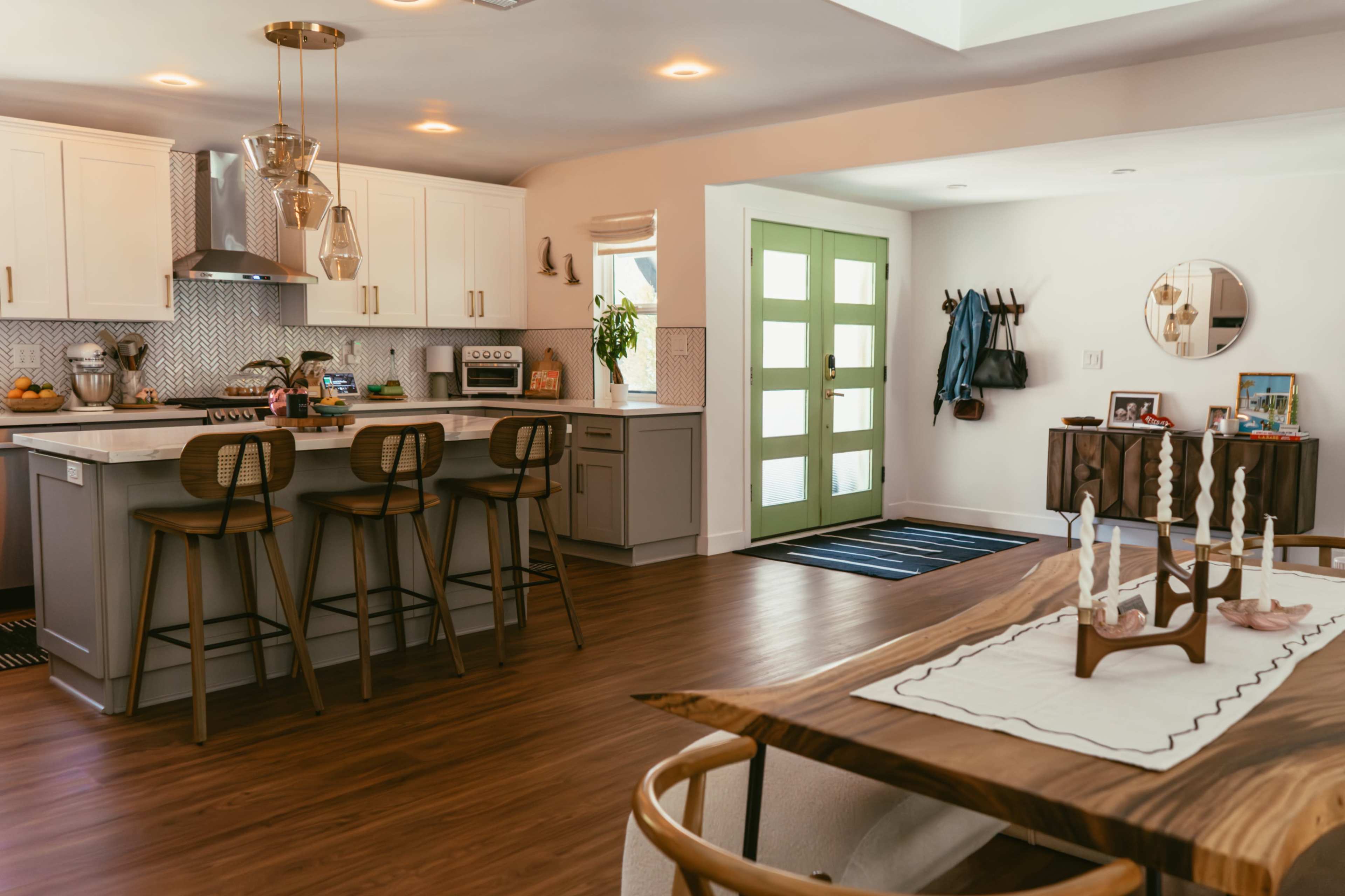 The image shows a modern kitchen with white cabinetry, a central island with bar stools, and an adjoining living area featuring a dining table and a green front door.