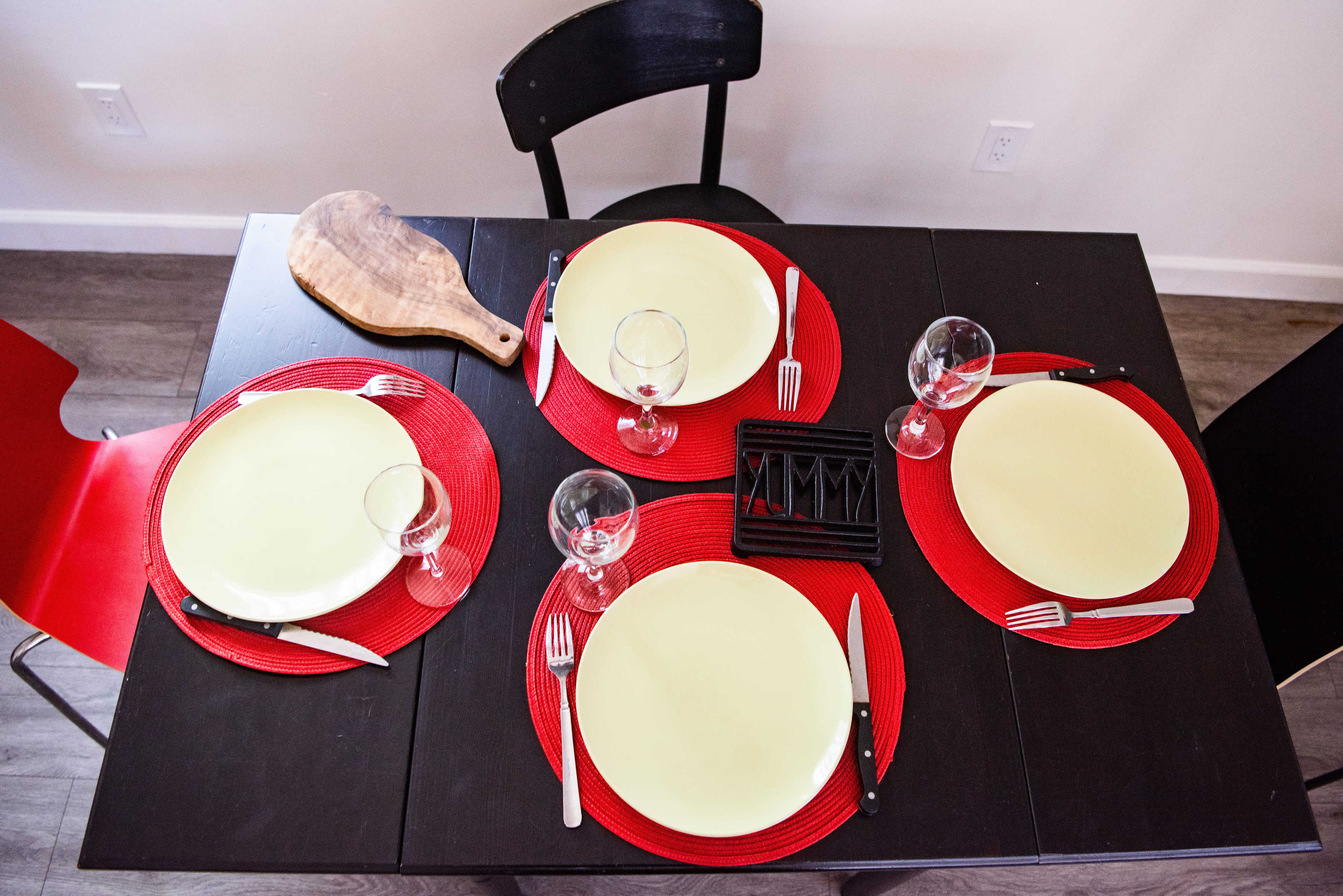 A dining table is set with four yellow plates on red placemats, accompanied by glasses and utensils, with a wooden cutting board nearby.