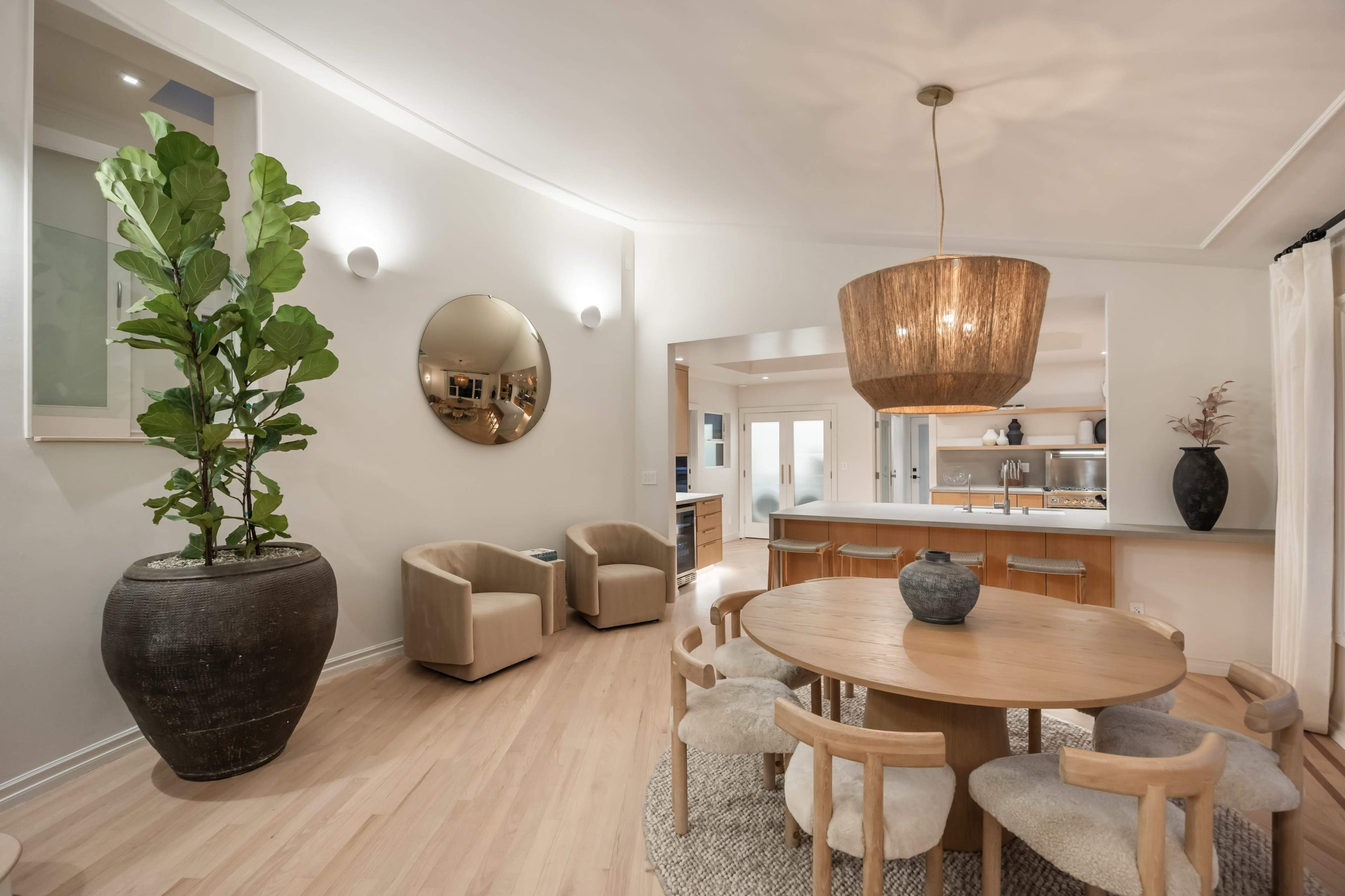 The image shows a modern interior space featuring a round wooden dining table surrounded by upholstered chairs, a large planter with a tall plant, and two stylish armchairs.