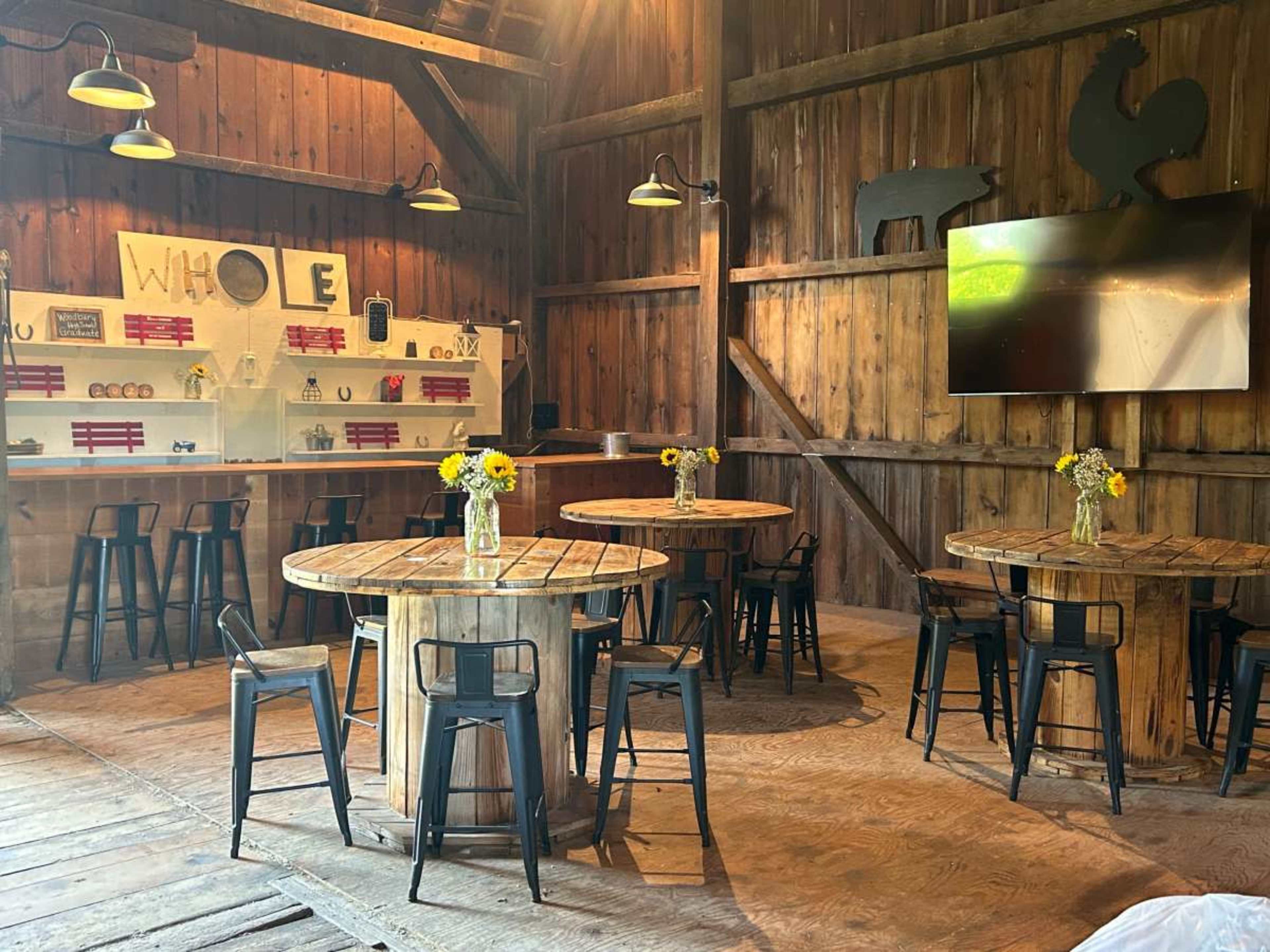 The image shows a rustic interior of a barn featuring wooden tables with black metal chairs, shelves displaying items, and yellow flowers in vases on the tables.