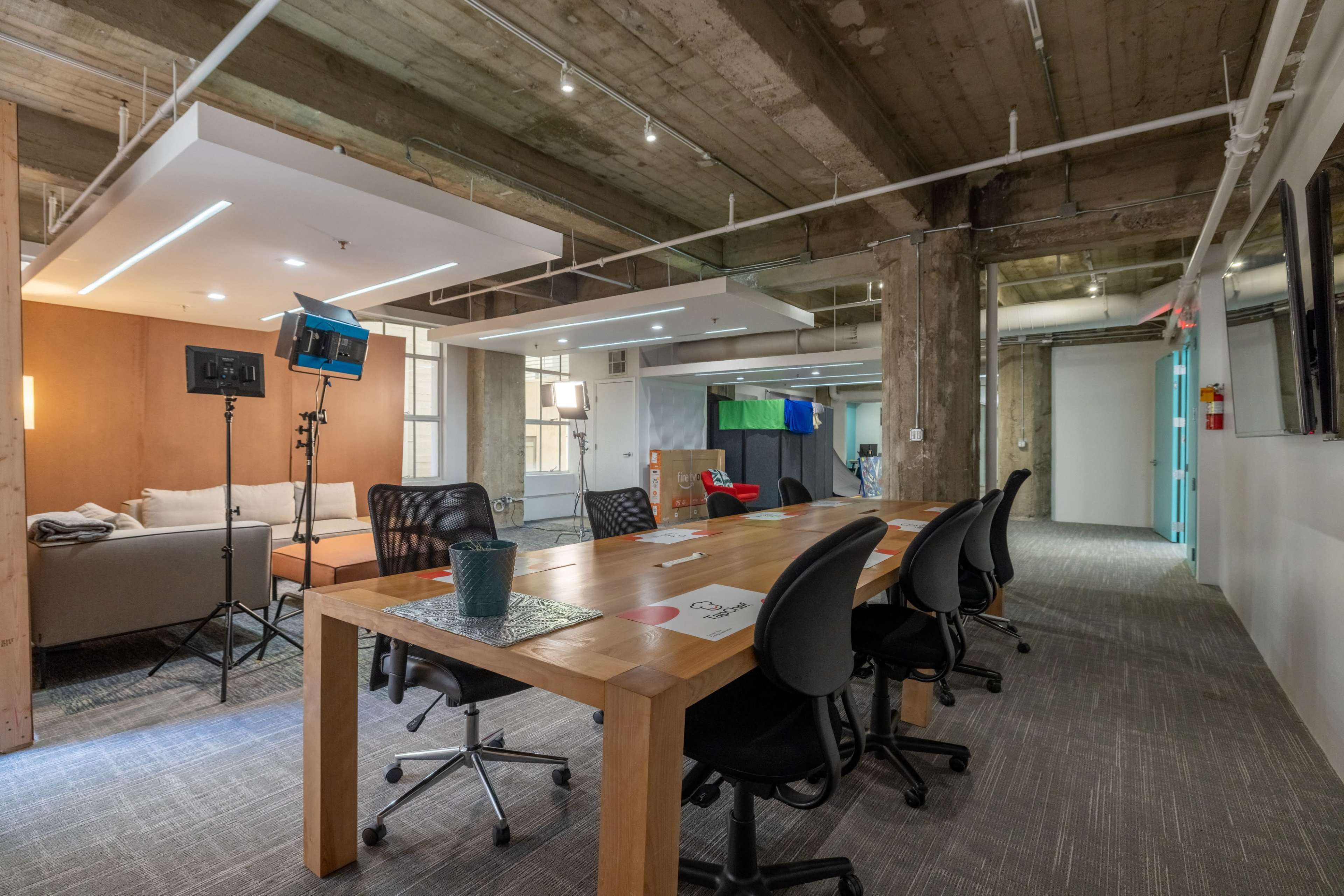The image shows a modern office space featuring a long wooden conference table surrounded by black chairs, with a lounge area and professional lighting equipment in the background.