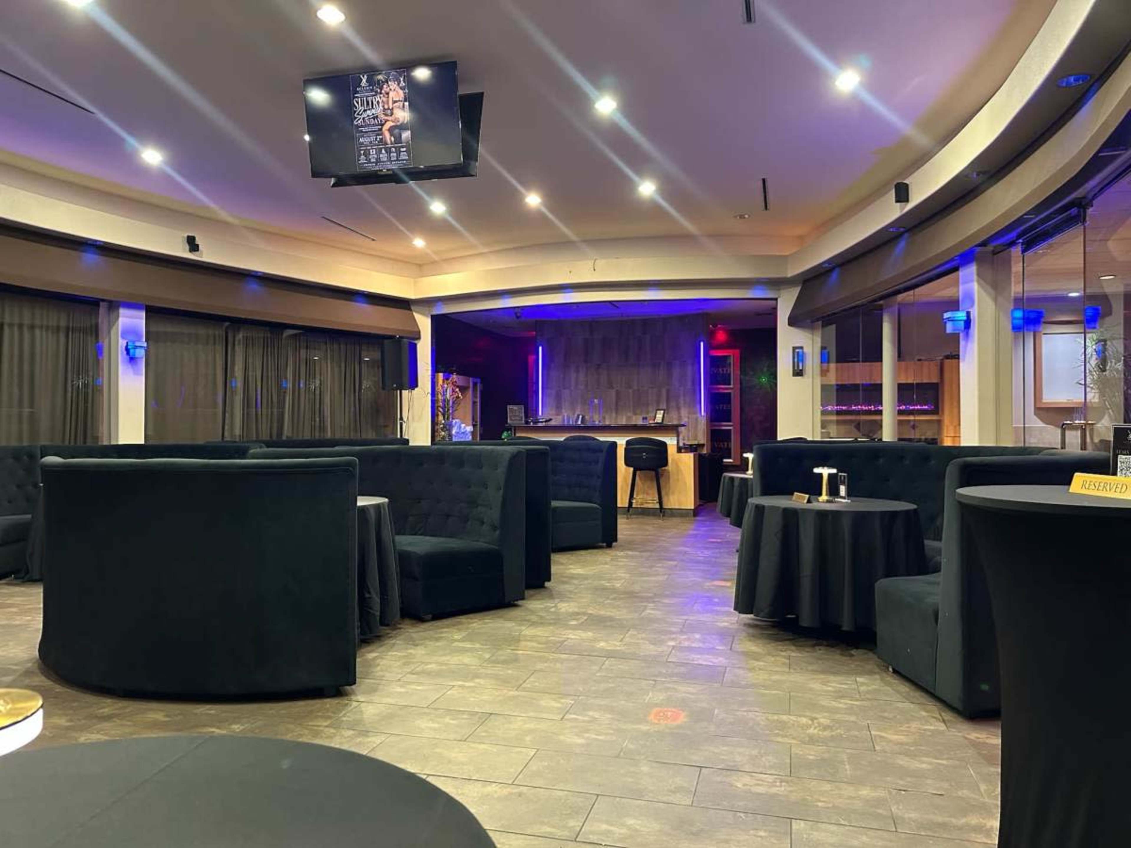 The image depicts an upscale lounge area with dark upholstered seating, circular tables, and ambient lighting, featuring a bar in the background.