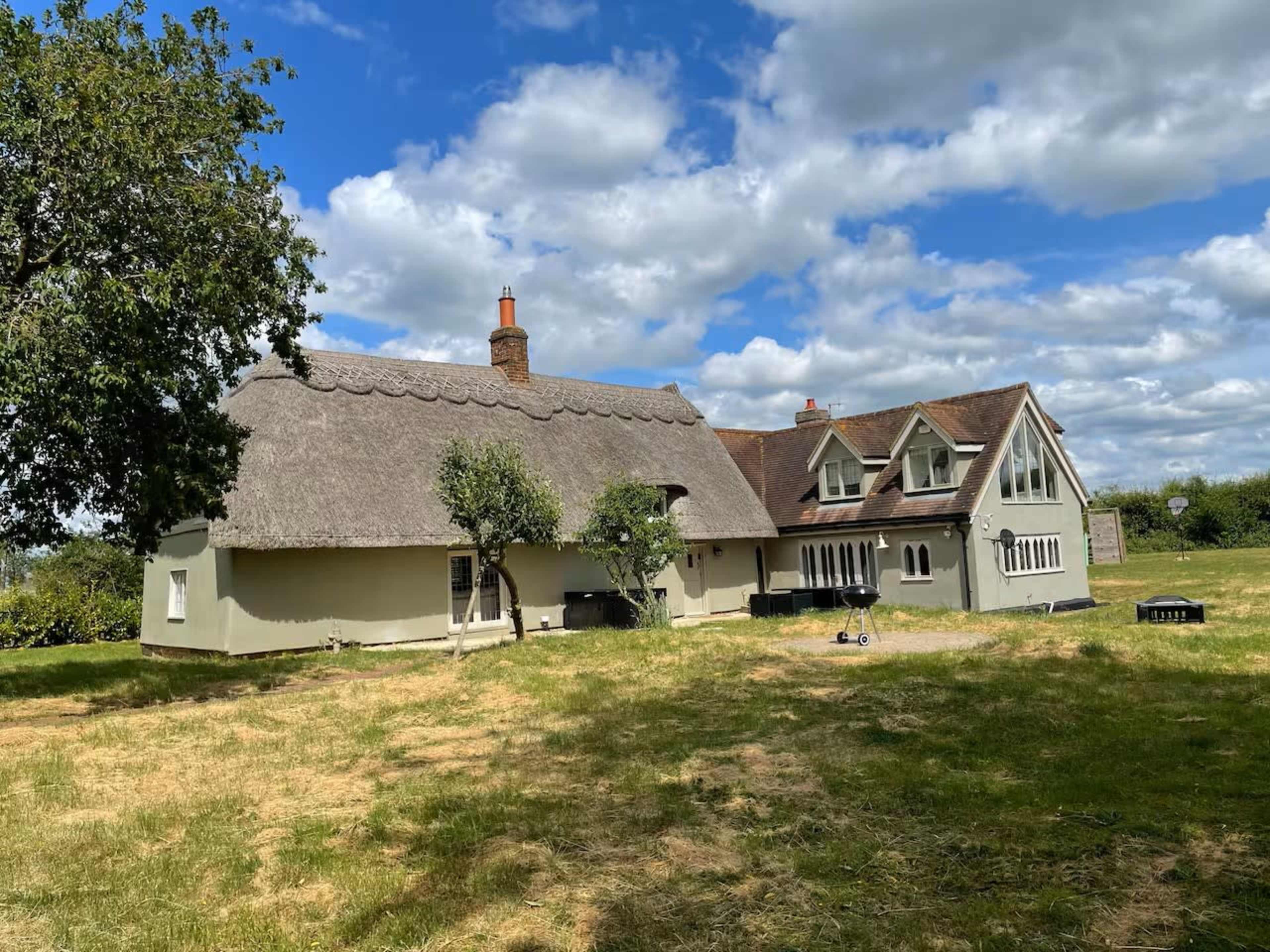 The image shows a two-part building, one with a thatched roof and the other with a modern design, set on a grassy area under a partly cloudy sky.
