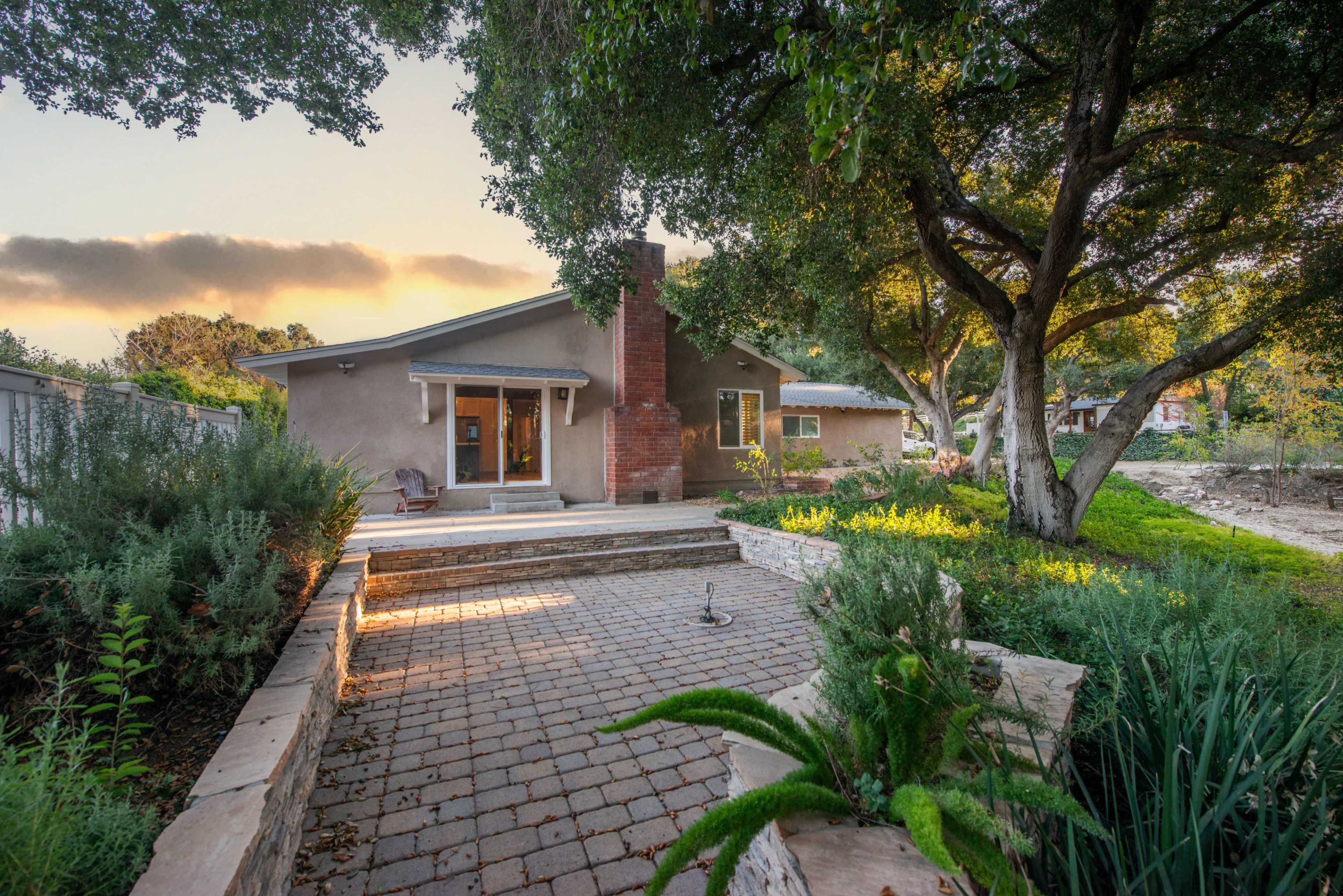 A backyard pathway leading to a single-story house surrounded by trees and landscaped greenery.