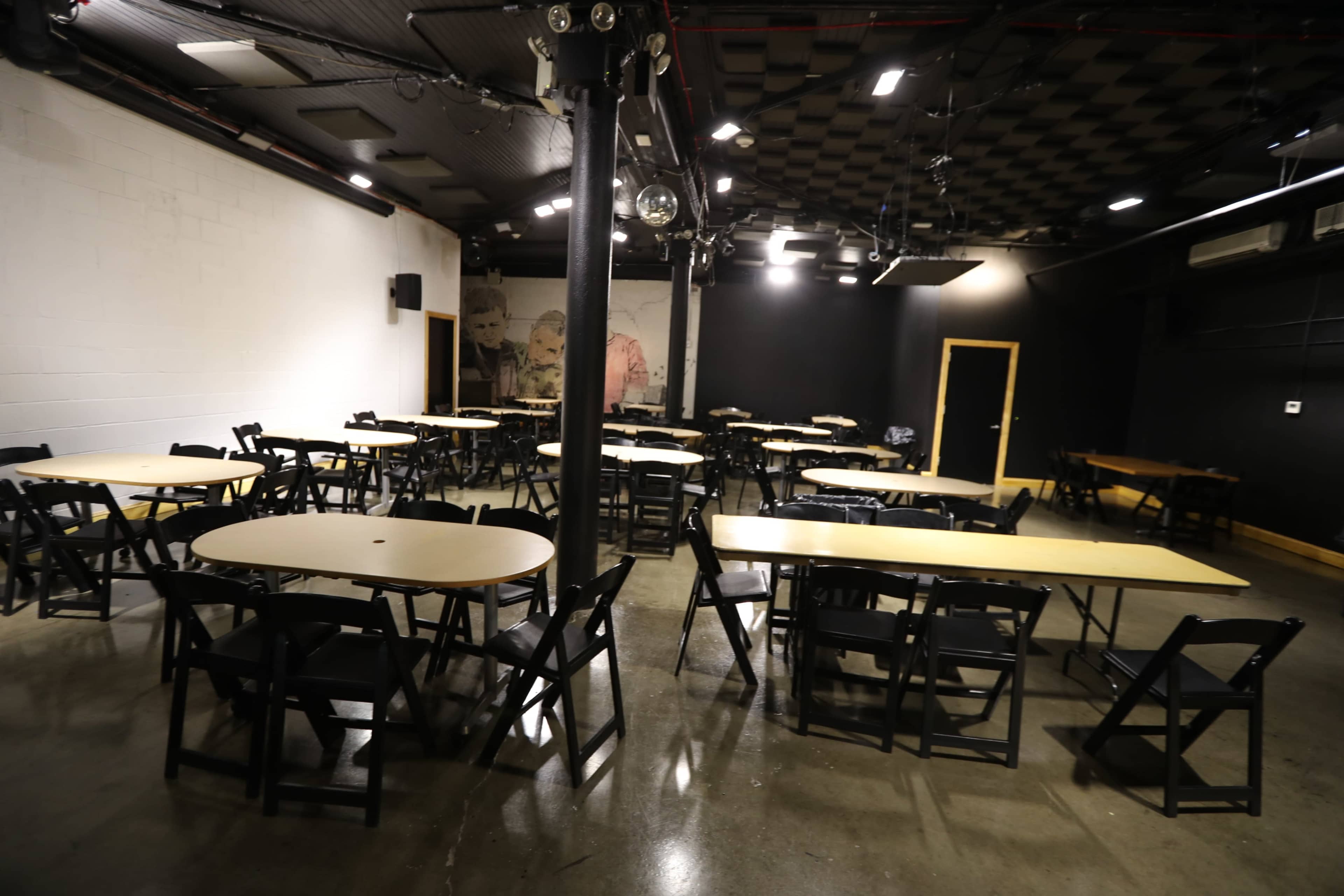 The image shows a spacious room with multiple round tables and black folding chairs arranged throughout, illuminated by overhead lights.