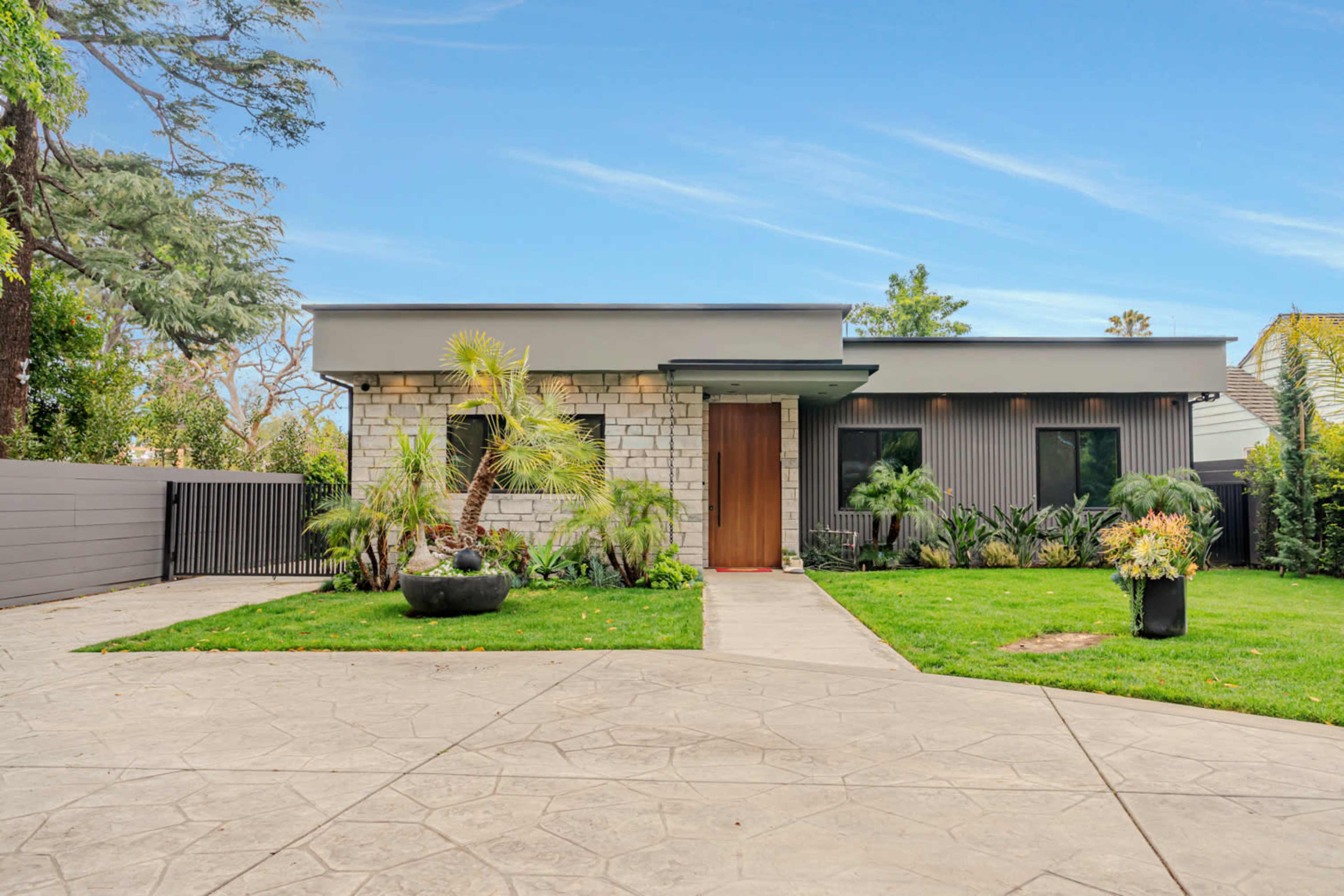 A modern single-story house with a stone and metal exterior, surrounded by a well-maintained lawn and diverse landscaping.