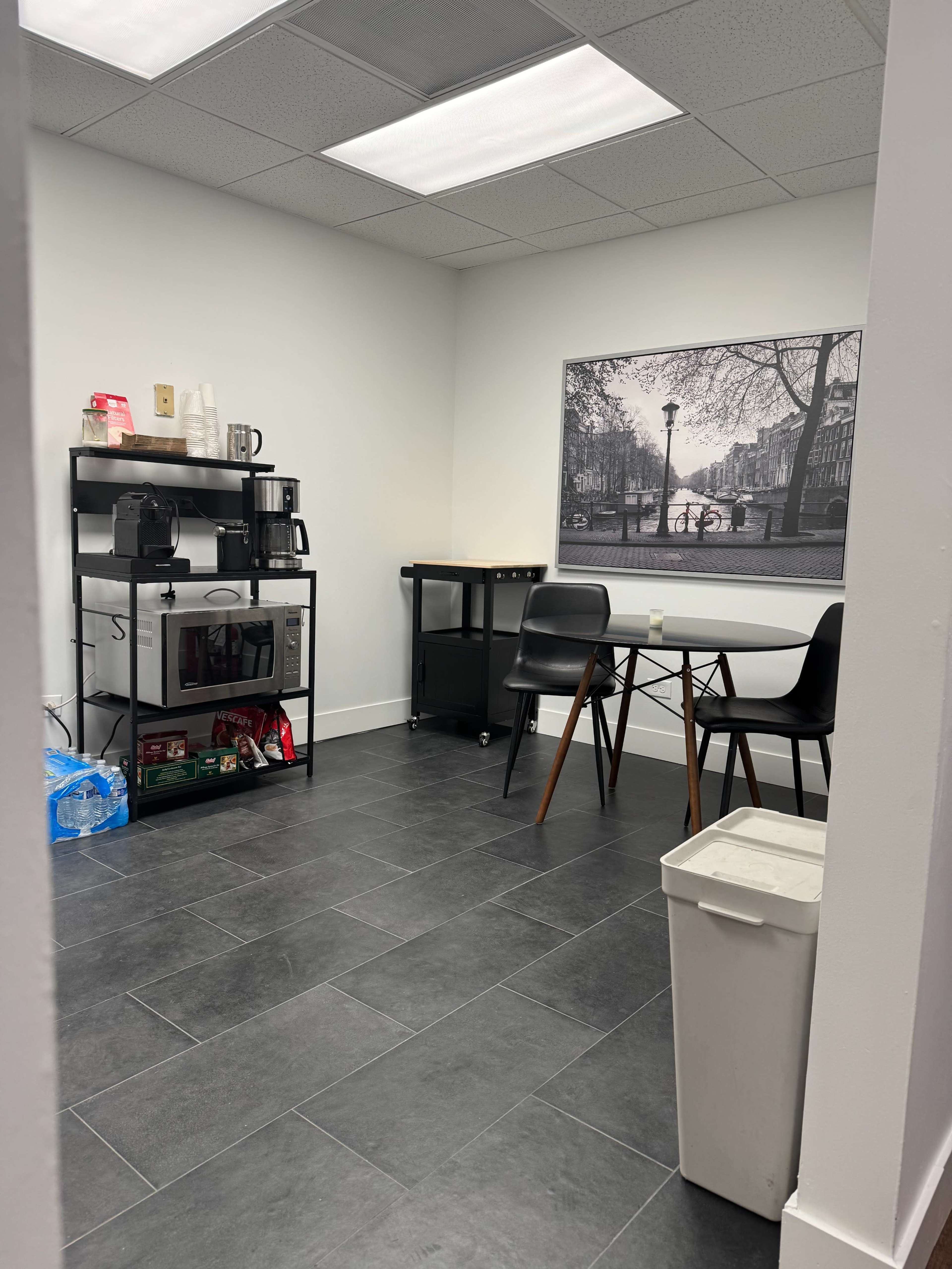 The image depicts a small break room with coffee-making equipment, a cart, a table with chairs, and a framed black-and-white photograph on the wall.