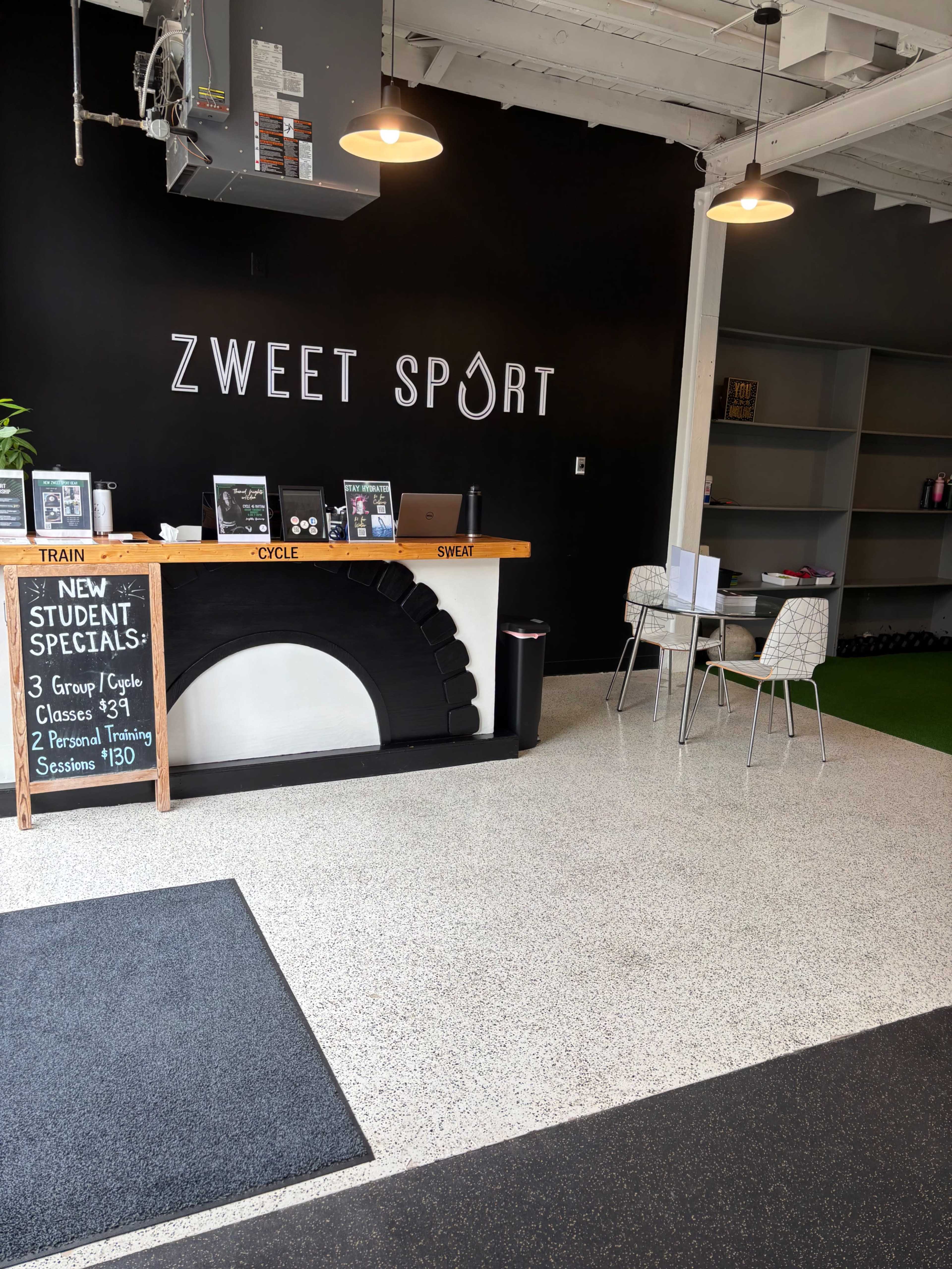The image shows a modern fitness studio reception area with a counter displaying information about classes and training sessions, and a black wall with the words "ZWEET SPORT" prominently featured.