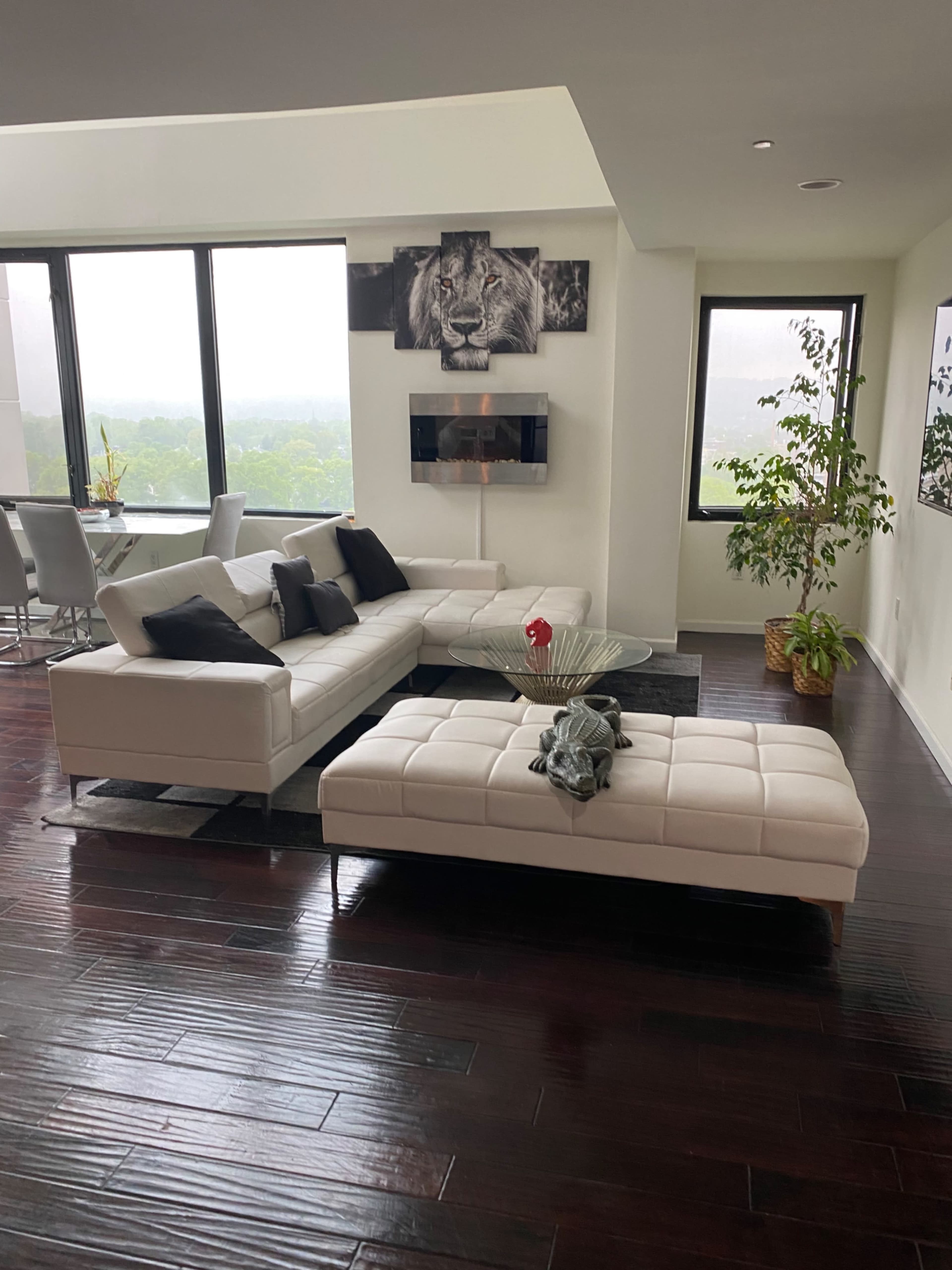 A modern living room with a white sectional sofa, a glass coffee table, and large windows offering a view of greenery outside.