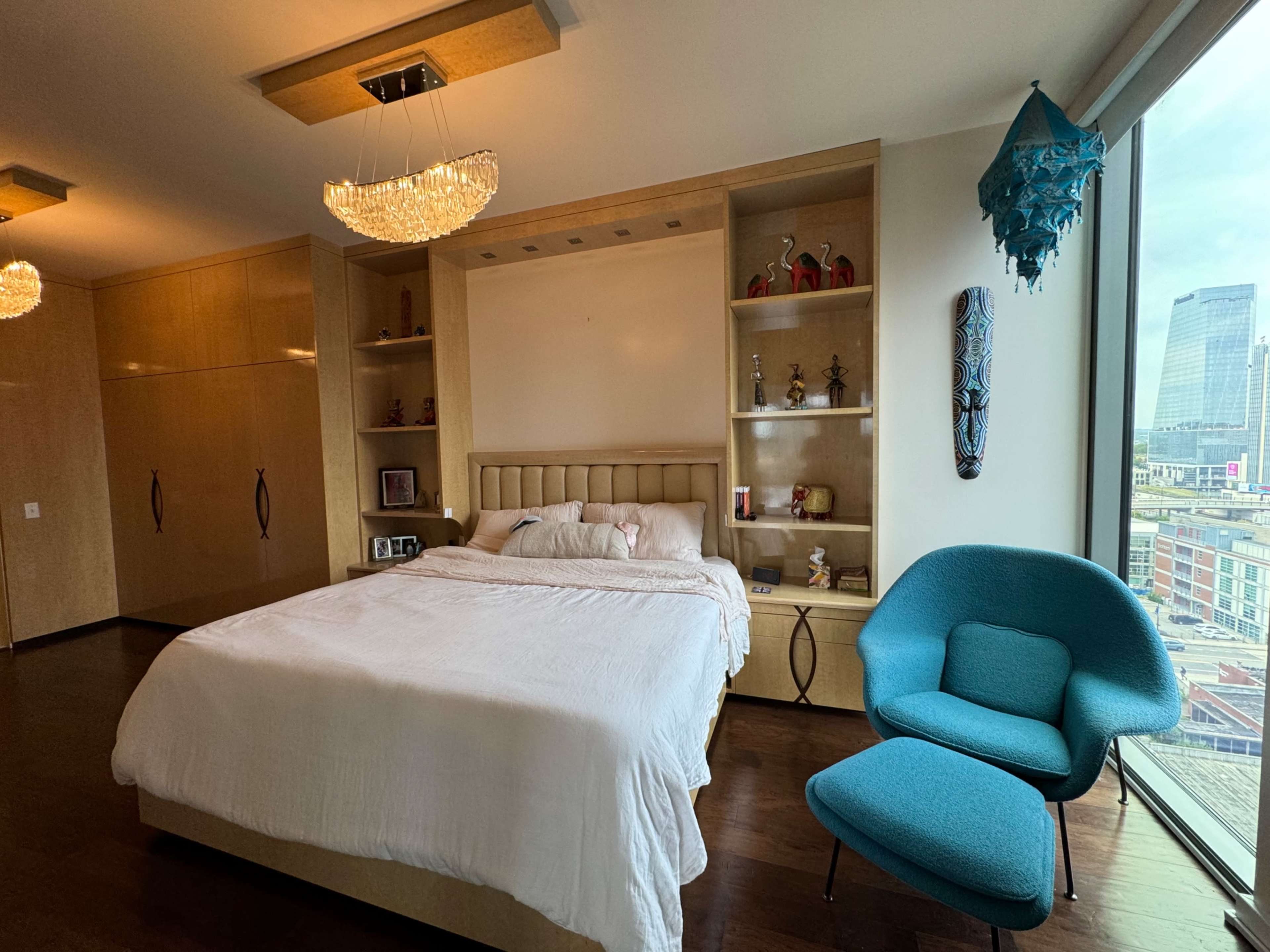 The room features a bed with white linens, a blue chair, and shelves displaying decorative items next to large windows.