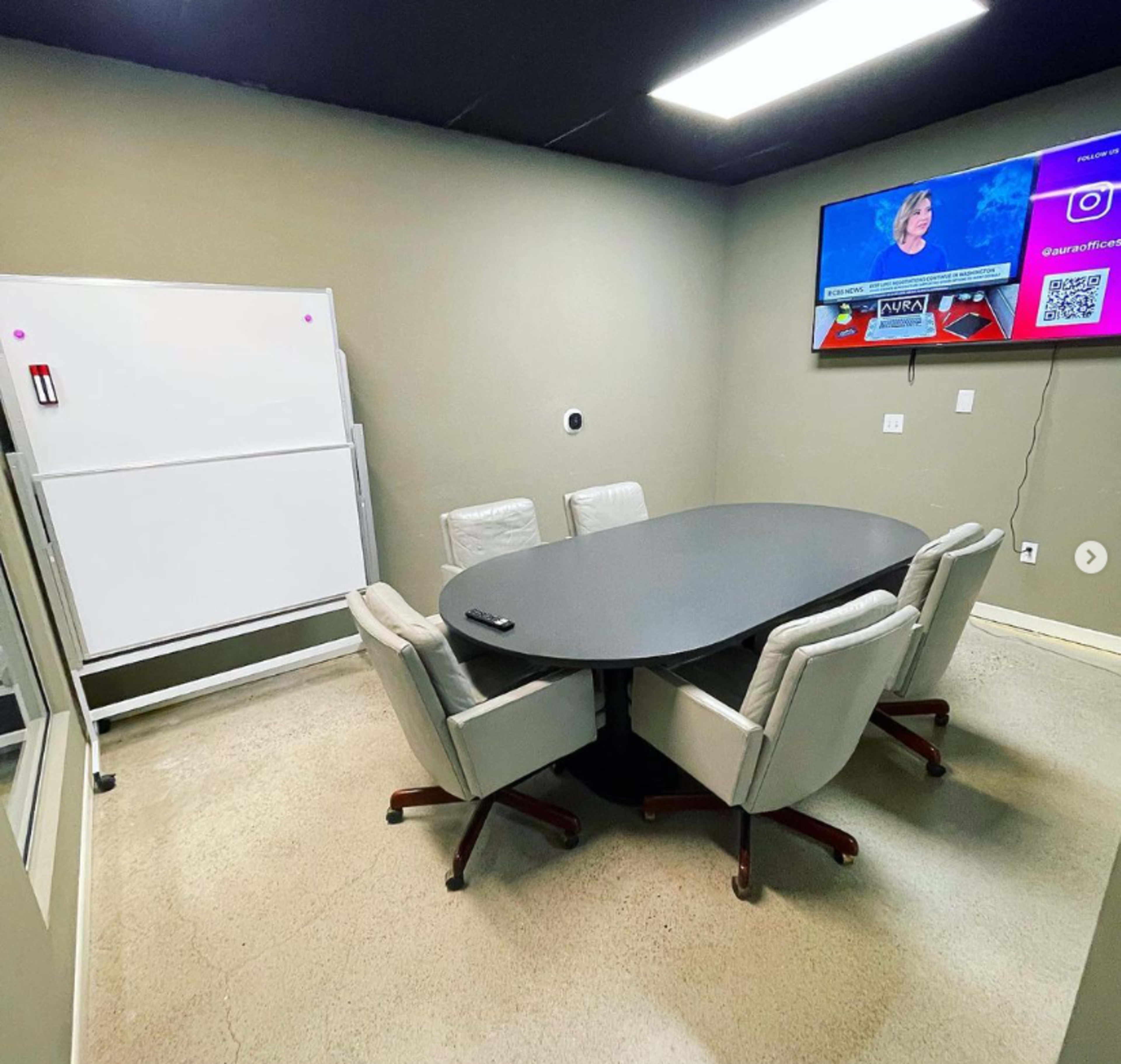 A conference room features a round table surrounded by four chairs, a whiteboard, and a television displaying news content.