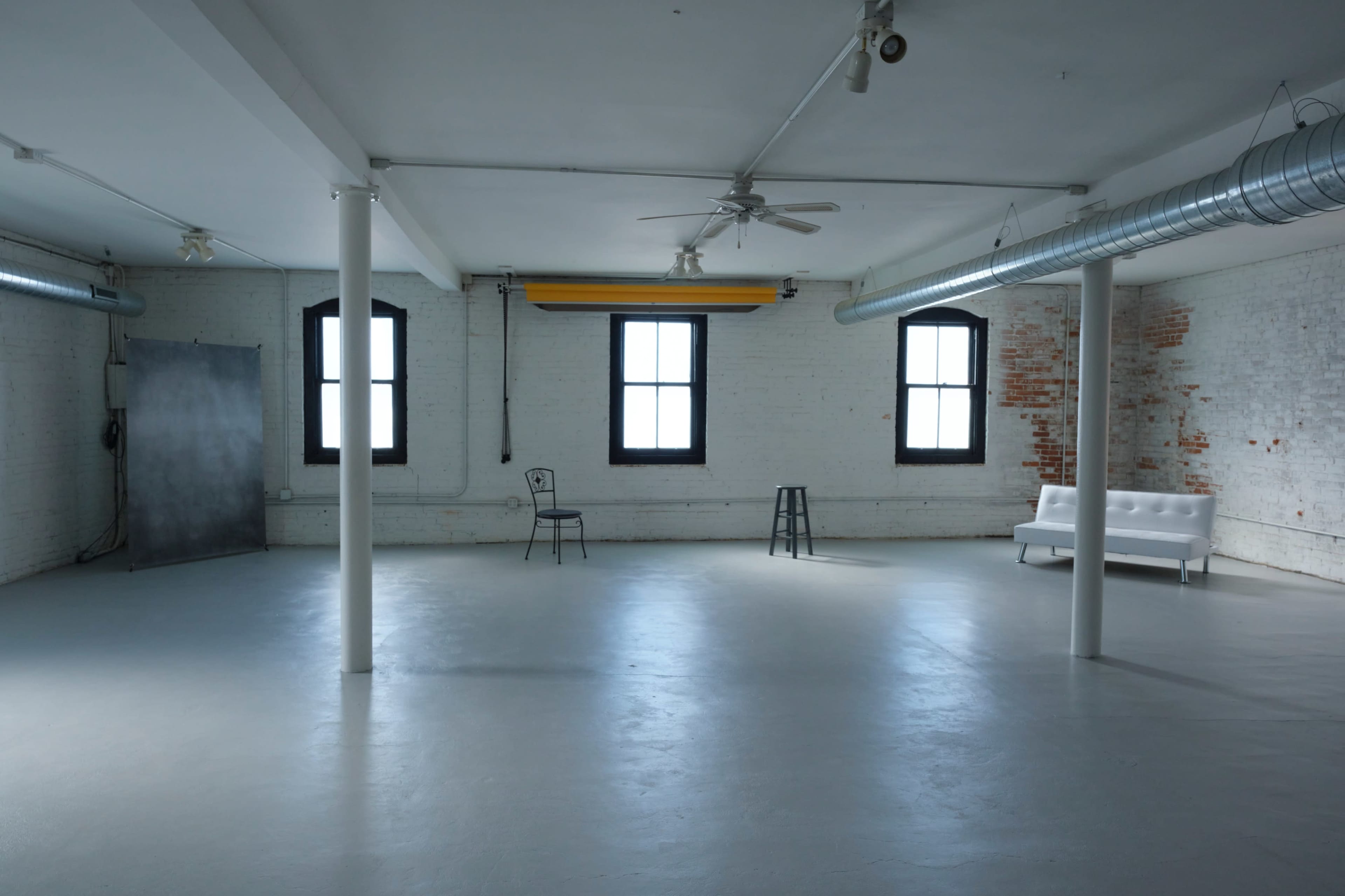 An empty industrial space features white walls, concrete flooring, two windows, a ceiling fan, and a white couch beside two stools.